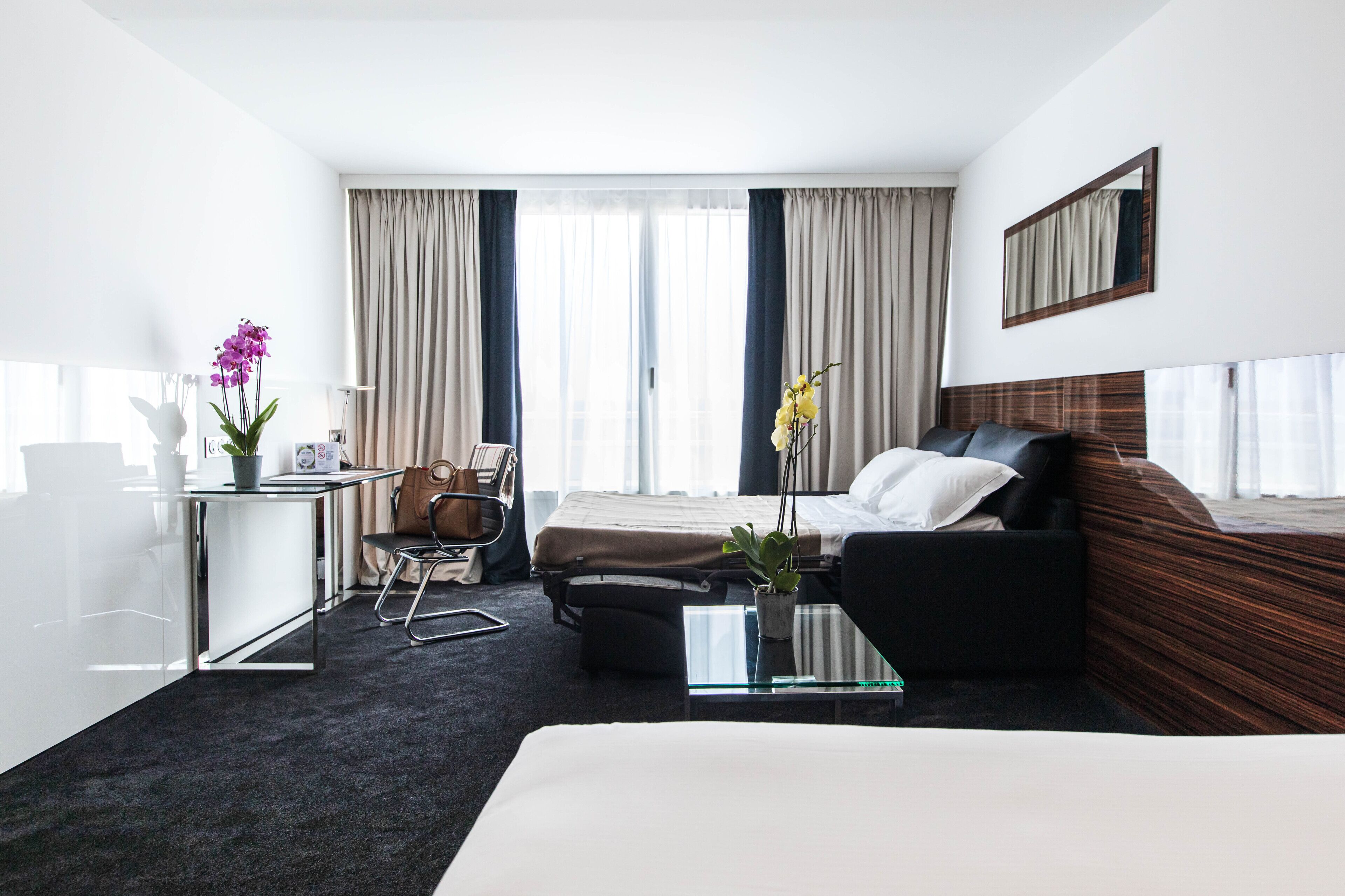 Une chambre d’hôtel avec un lit, un canapé, un bureau et une chaise. Il y a des rideaux, un miroir et une fenêtre.
