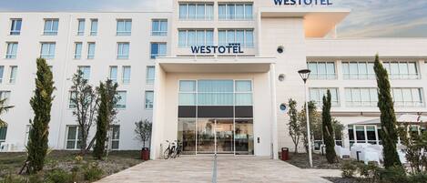 Un edificio Westotel con diseño moderno, que cuenta con una entrada amplia, múltiples ventanas y un área exterior bien cuidada con plantas en macetas y un camino.