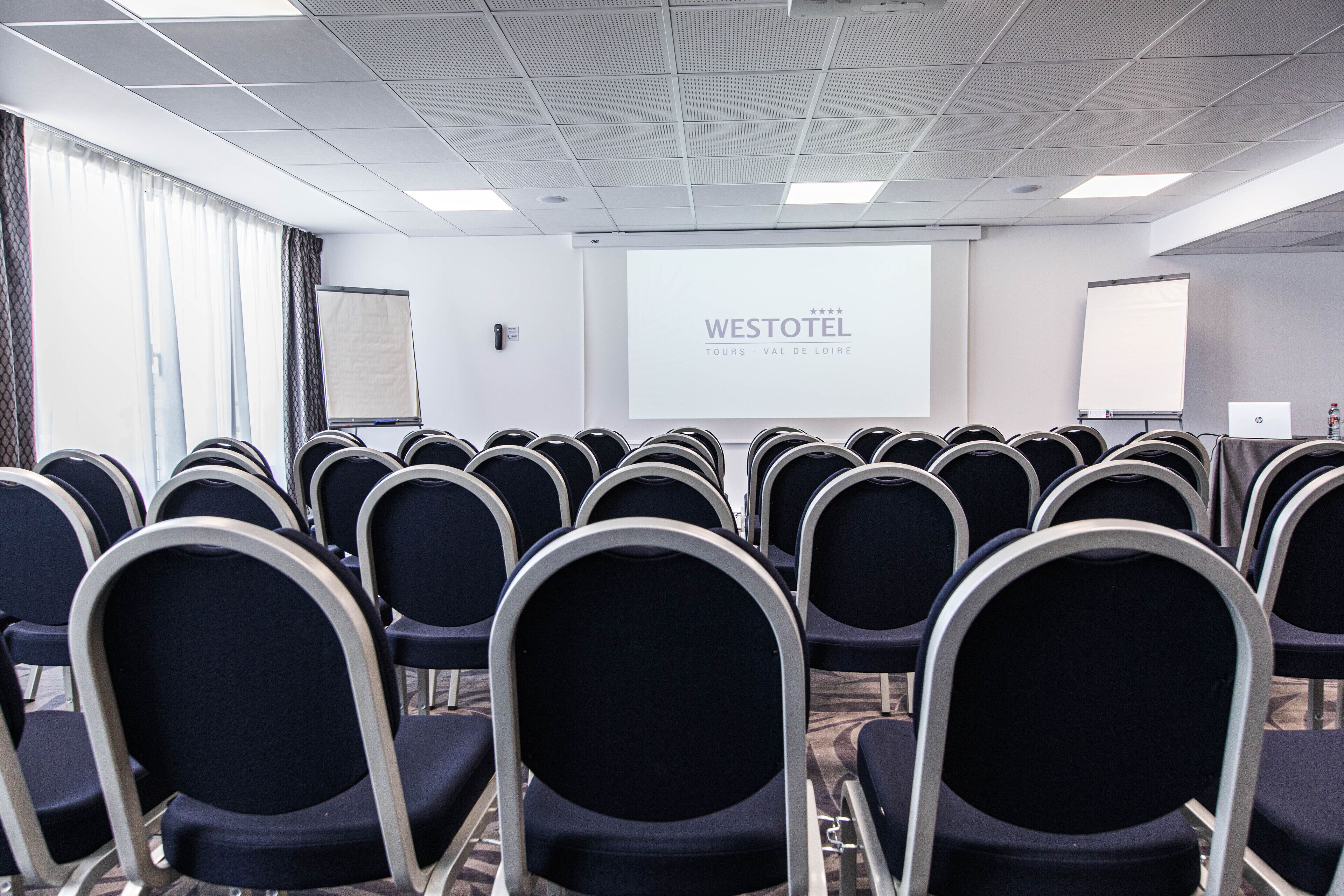 Une salle de conférence avec des rangées de chaises, un écran de projection affichant le texte « WESTOTEL TOURS - VAL DE LOIRE », et un tableau blanc.