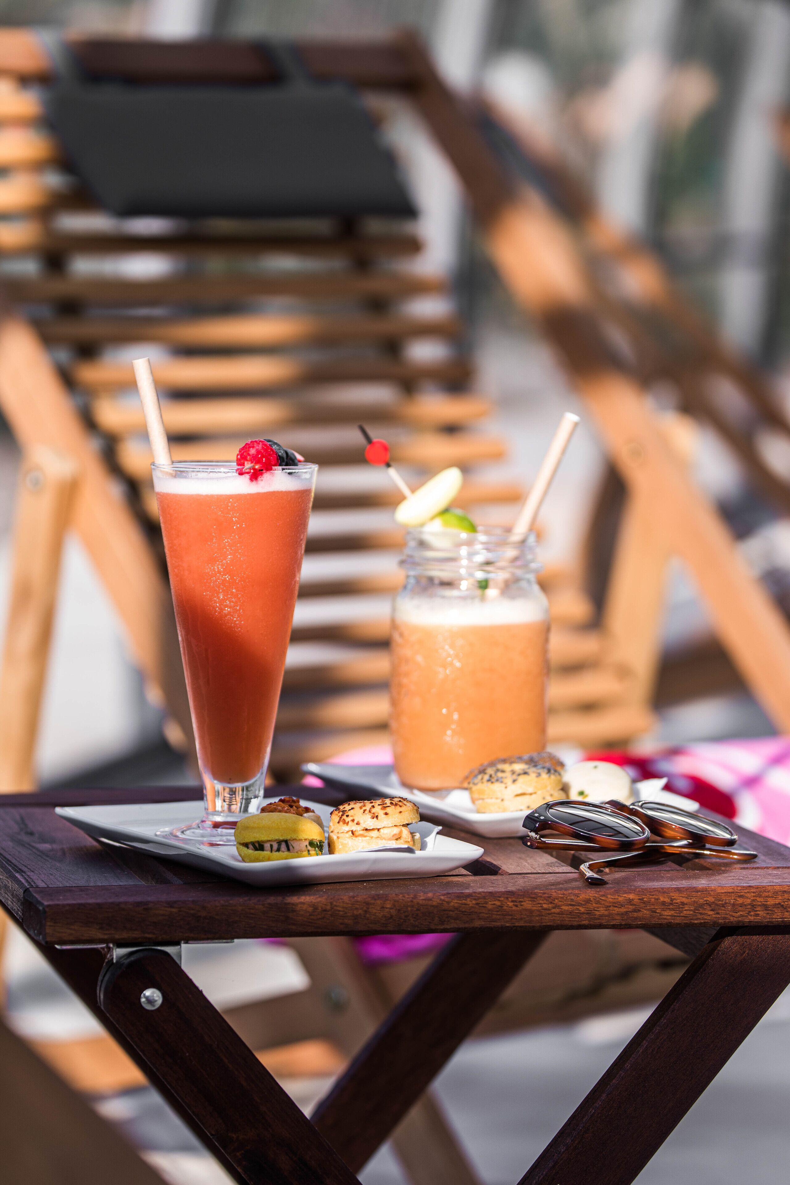A table with a tall glass of iced beverage garnished with a berry and a cherry, a mason jar with a straw, and a plate with food items.
