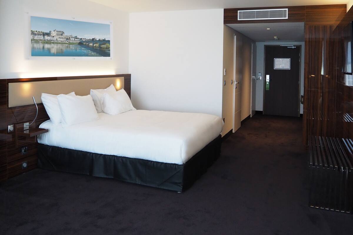 Ein Hotelzimmer mit einem großen Bett, einem Nachttisch, einem Wand-Fernseher und einer Tür zu einem Badezimmer.