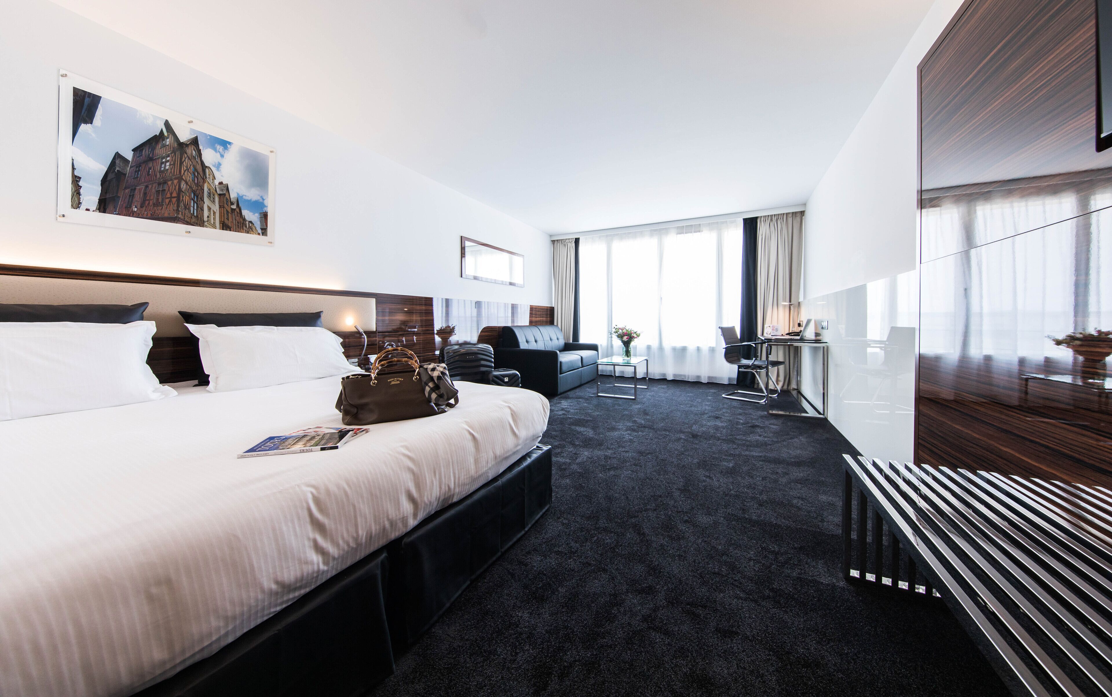 A modern hotel room with a large bed, a desk, a chair, a sofa, and a view of the city.