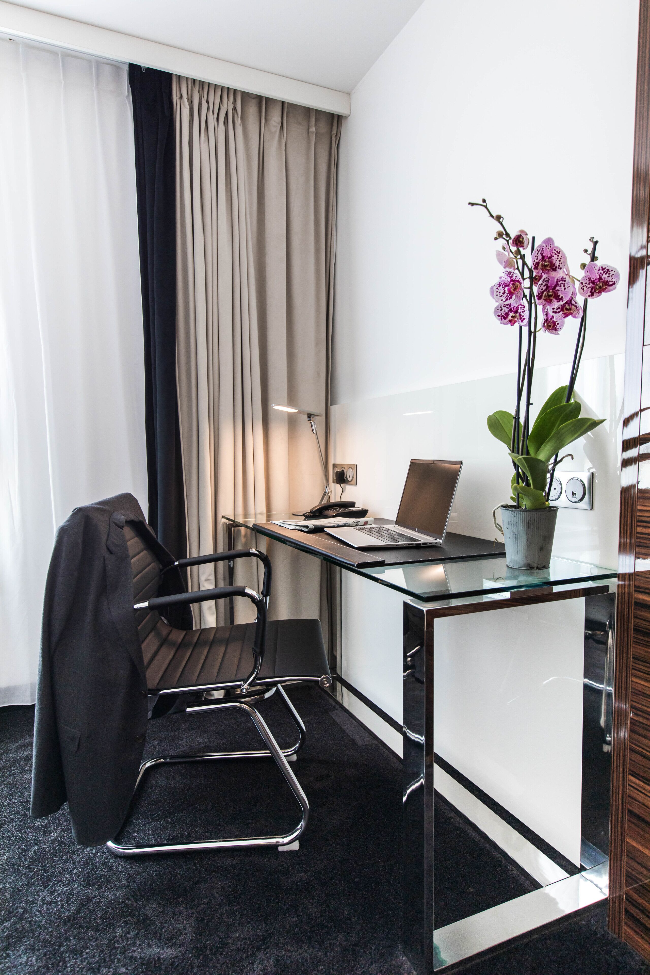 Een moderne hotelkamer met een glazen bureau, een laptop, een potorkidee, een zwarte stoel en een raam met gordijnen.