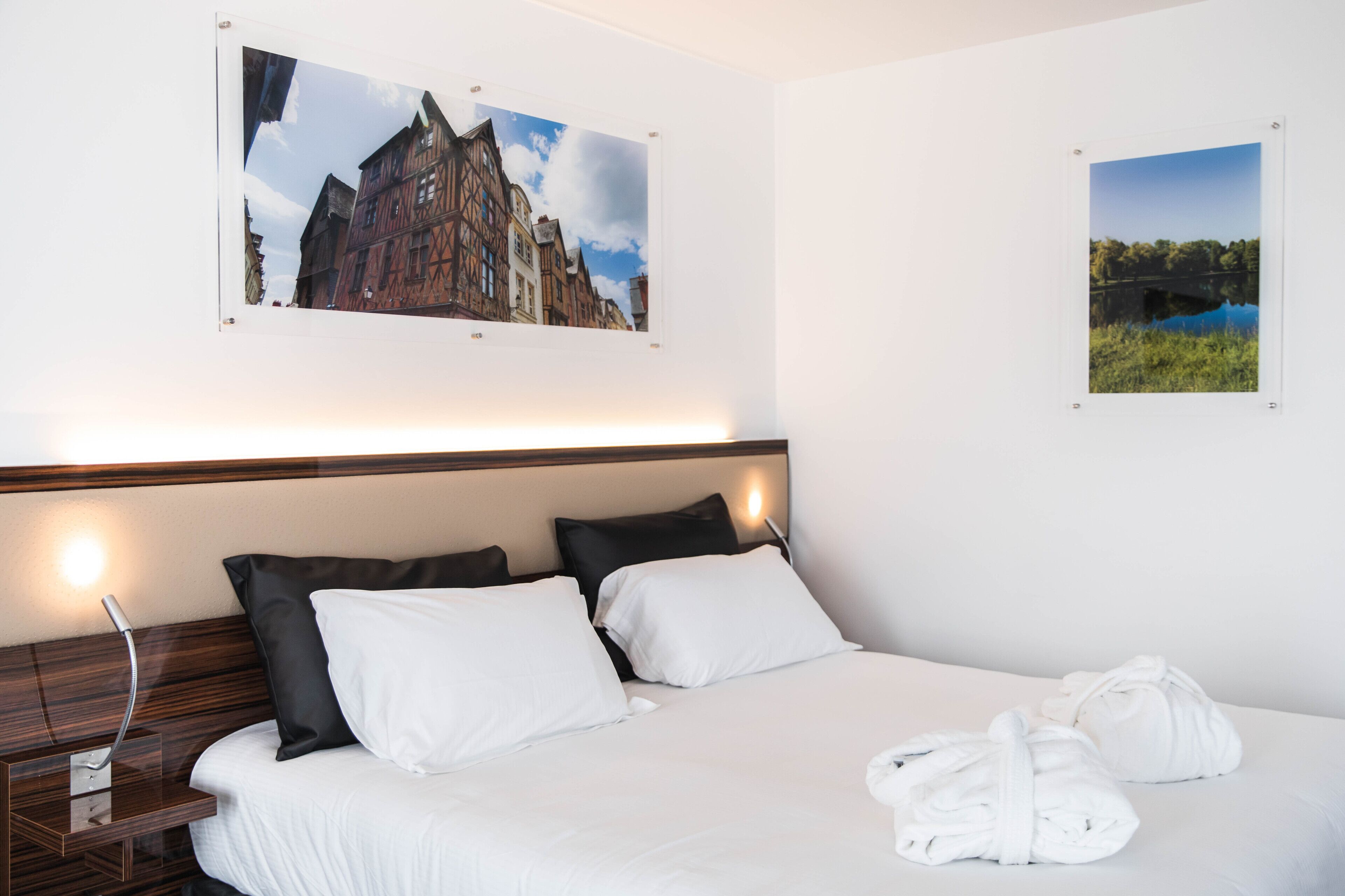 A hotel room with a large bed, two pillows, a bedside table with a lamp, and two framed pictures on the wall.