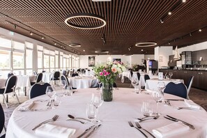Une salle de banquet avec des tables rondes dressées pour le repas, un arrangement floral central et un design de plafond moderne.
