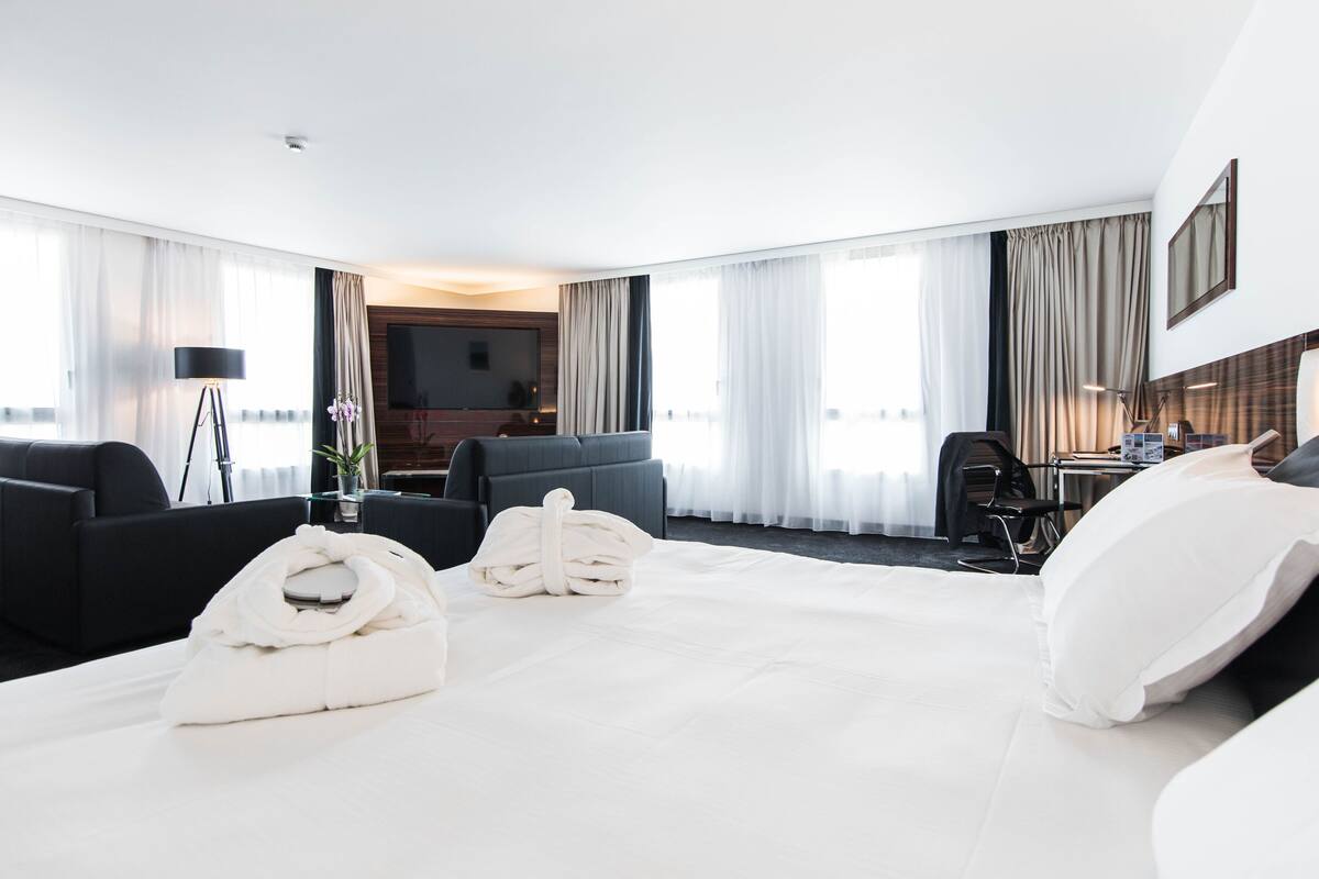 Ein modernes Hotelzimmer mit einem großen Bett, zwei Sesseln, einem Schreibtisch mit Stuhl und einem Fernseher.