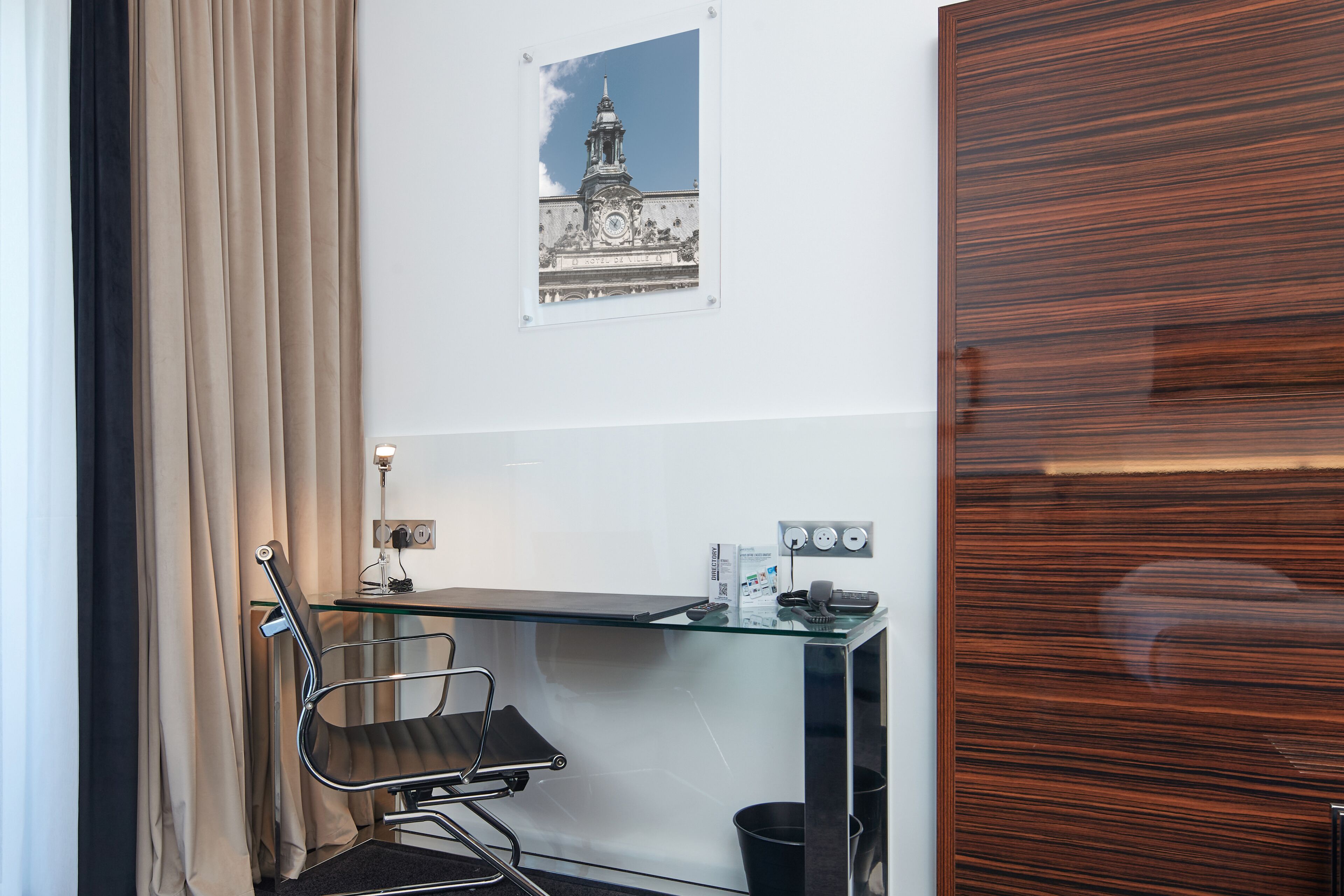 A modern hotel room with a glass desk, a black office chair, a black trash bin, and a framed picture of a building on the wall.