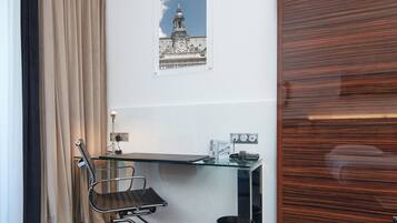Una habitación de hotel moderna con un escritorio de vidrio, una silla de oficina negra, un cesto de basura negro y un cuadro enmarcado de un edificio en la pared.