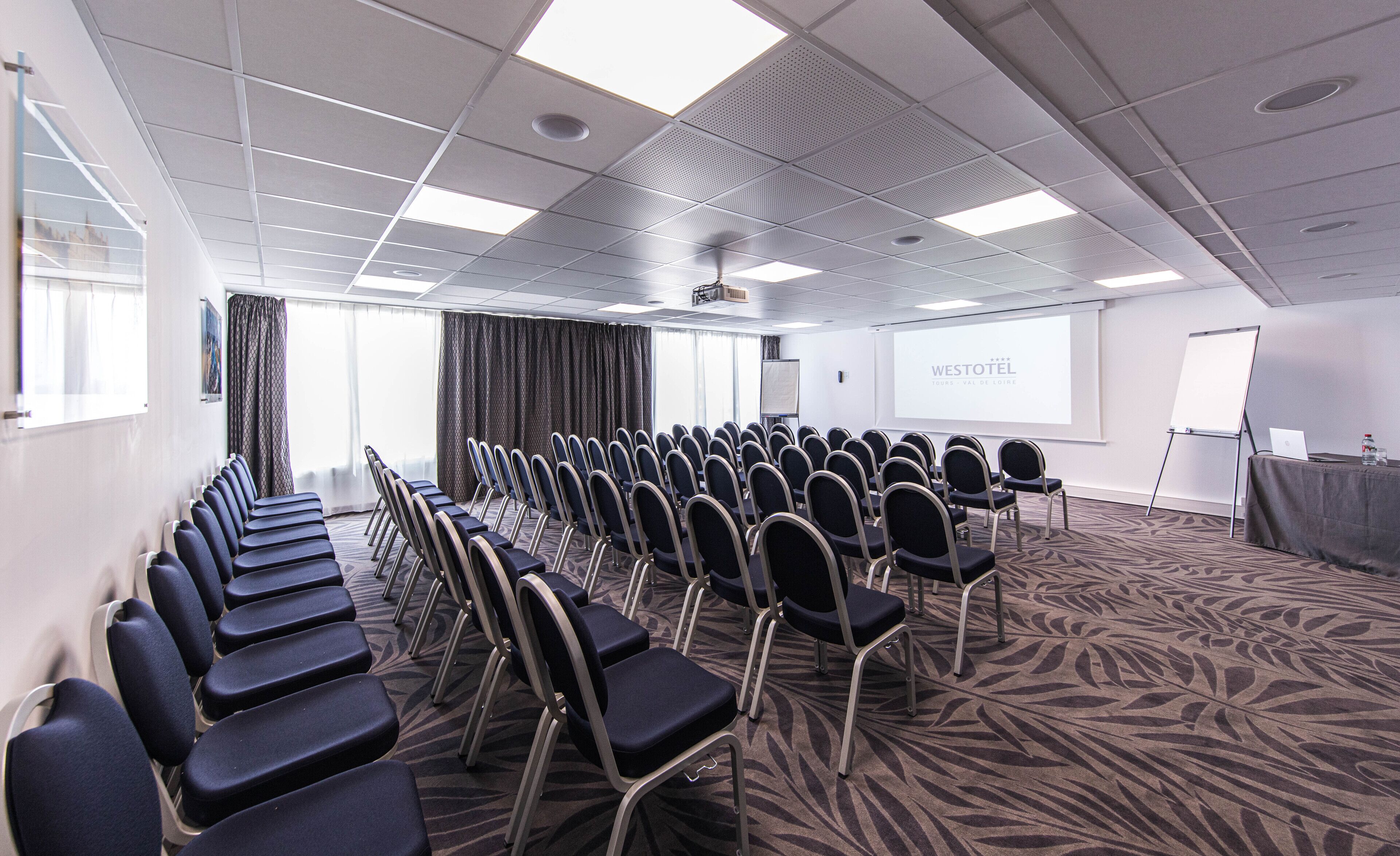 Un salón de conferencias con filas de sillas, una pantalla de proyector y una pizarra.