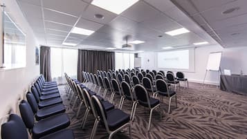 Uma sala de conferências com fileiras de cadeiras, uma tela de projetor e uma lousa.