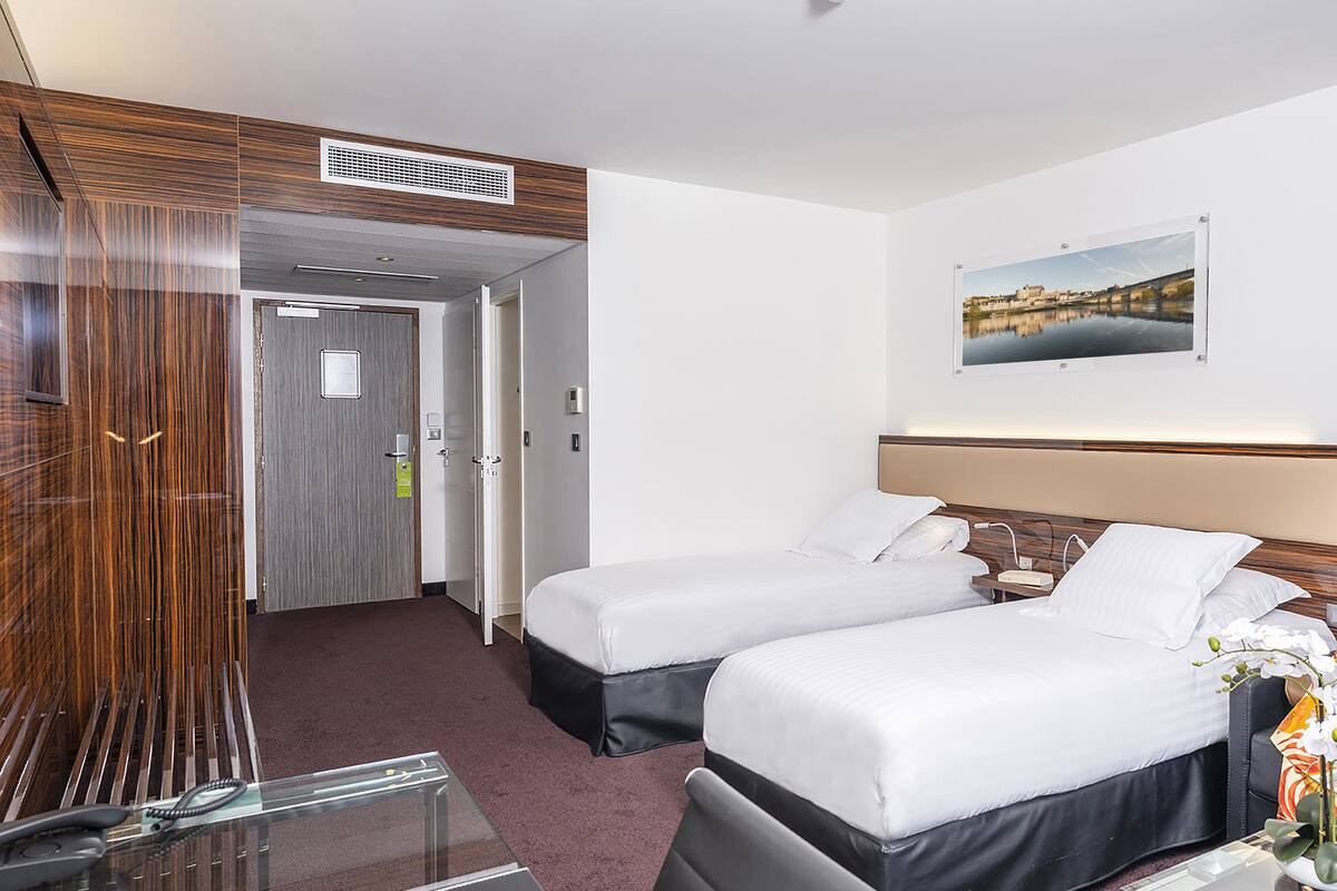 Ein Hotelzimmer mit zwei Betten, einem Schreibtisch mit Telefon, einem Bild an der Wand und einer Tür zu einem weiteren Zimmer.