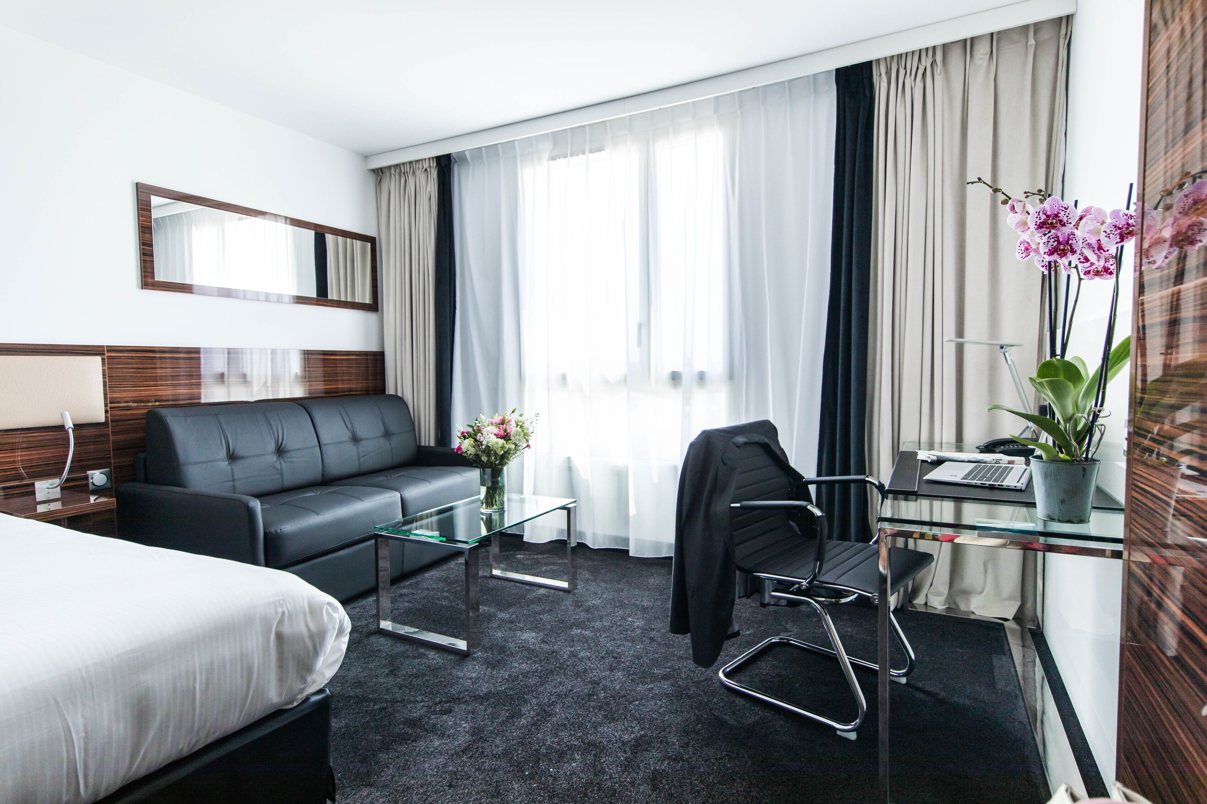 A hotel room with a bed, a black leather sofa, a glass coffee table, a desk with a laptop, a chair, and a potted plant.