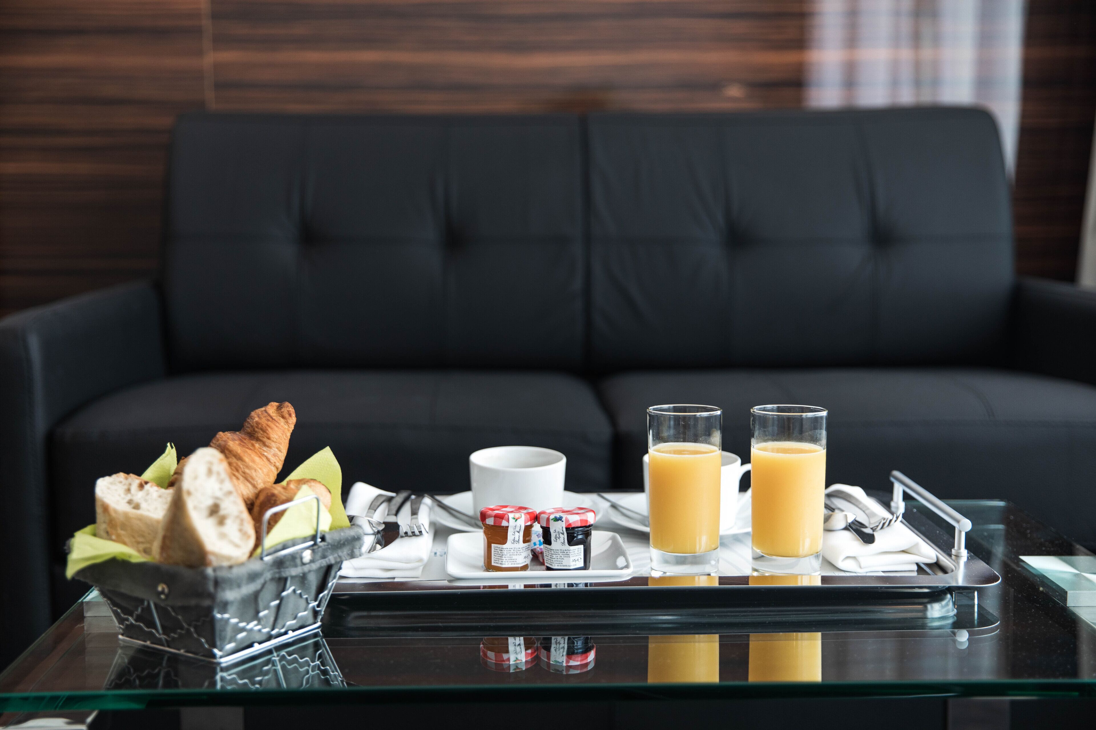 En bakke med croissanter, marmelade, juice og en kop på et glastop i forgrunden af en sort sofa.