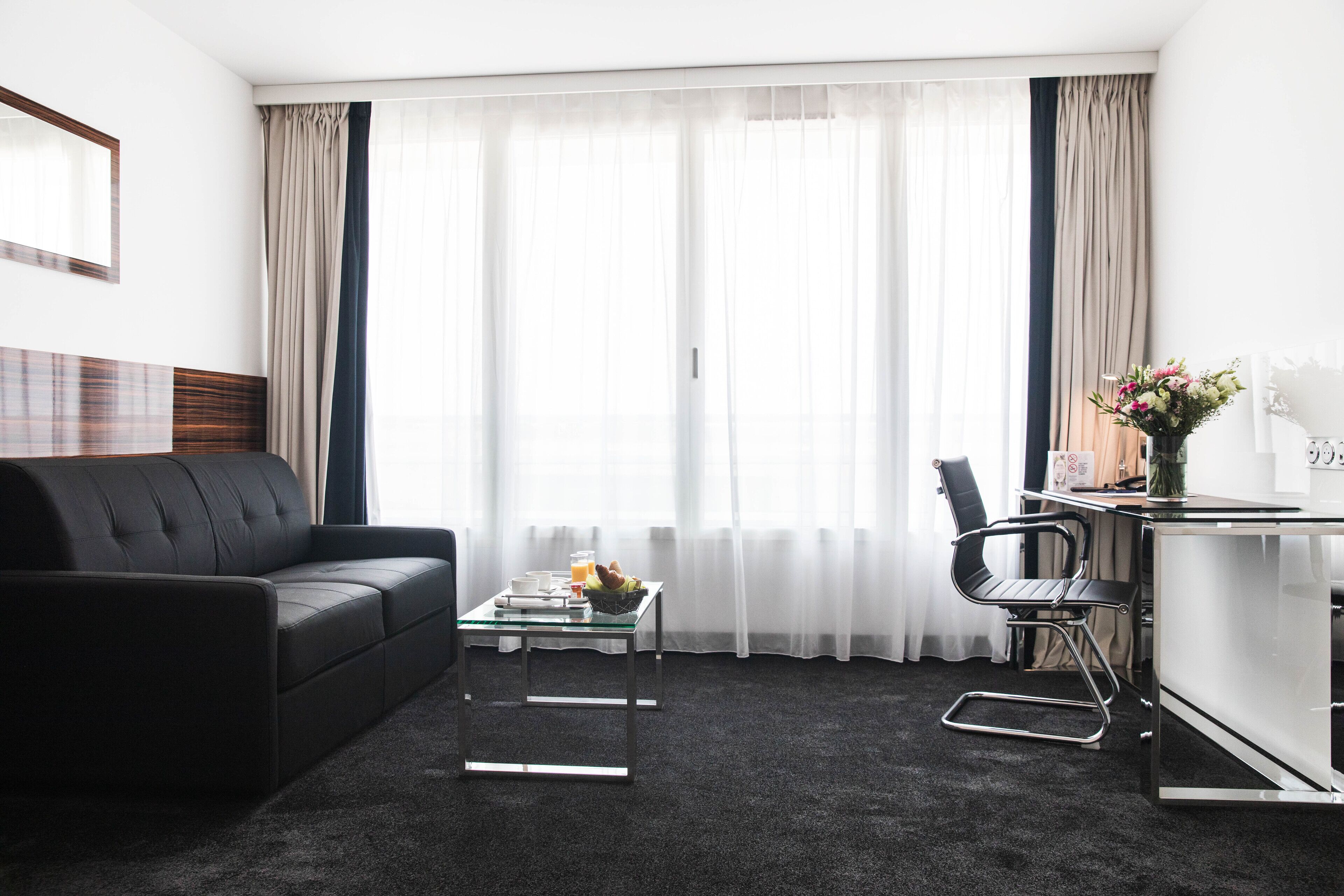 Une chambre d’hôtel moderne avec un canapé en cuir noir, une table basse en verre, un fauteuil de bureau noir et un petit bureau agrémenté d’un vase de fleurs.