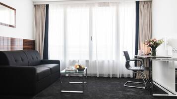 Une chambre d’hôtel moderne avec un canapé en cuir noir, une table basse en verre, un fauteuil de bureau noir et un petit bureau agrémenté d’un vase de fleurs.