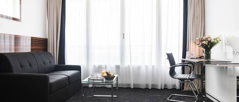 A modern hotel room with a black leather sofa, a glass coffee table, a black office chair, and a small desk with a vase of flowers.