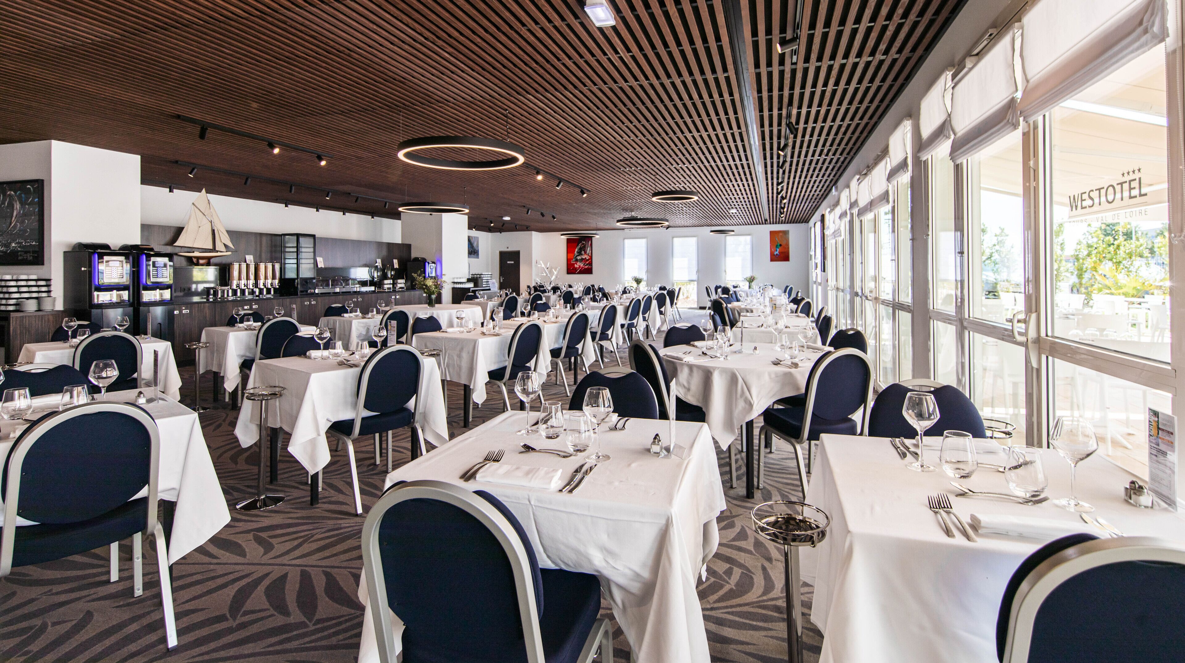 A spacious dining area with multiple tables set for guests, featuring a modern ceiling design and large windows offering a view of the outdoors.