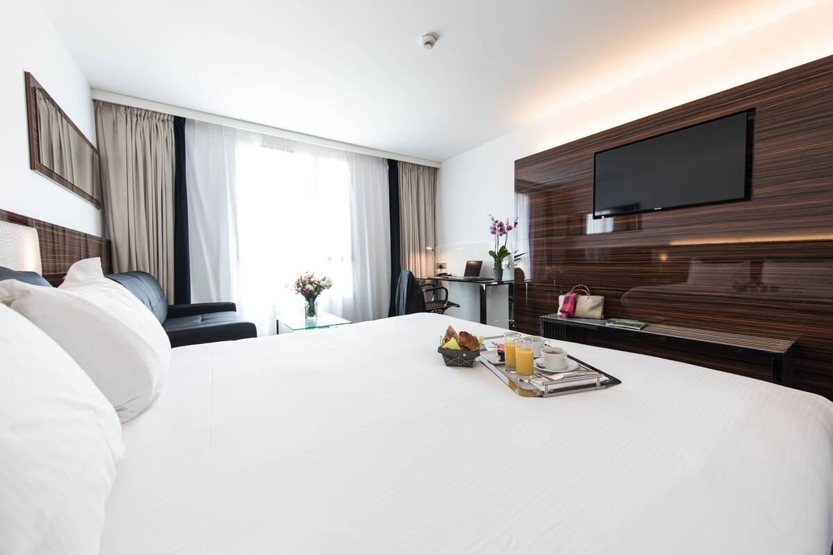 Ein Hotelzimmer mit einem großen Bett, einem flachbildfernseher an der Wand, einem Schreibtisch mit Stuhl und einem Fenster mit Vorhängen.