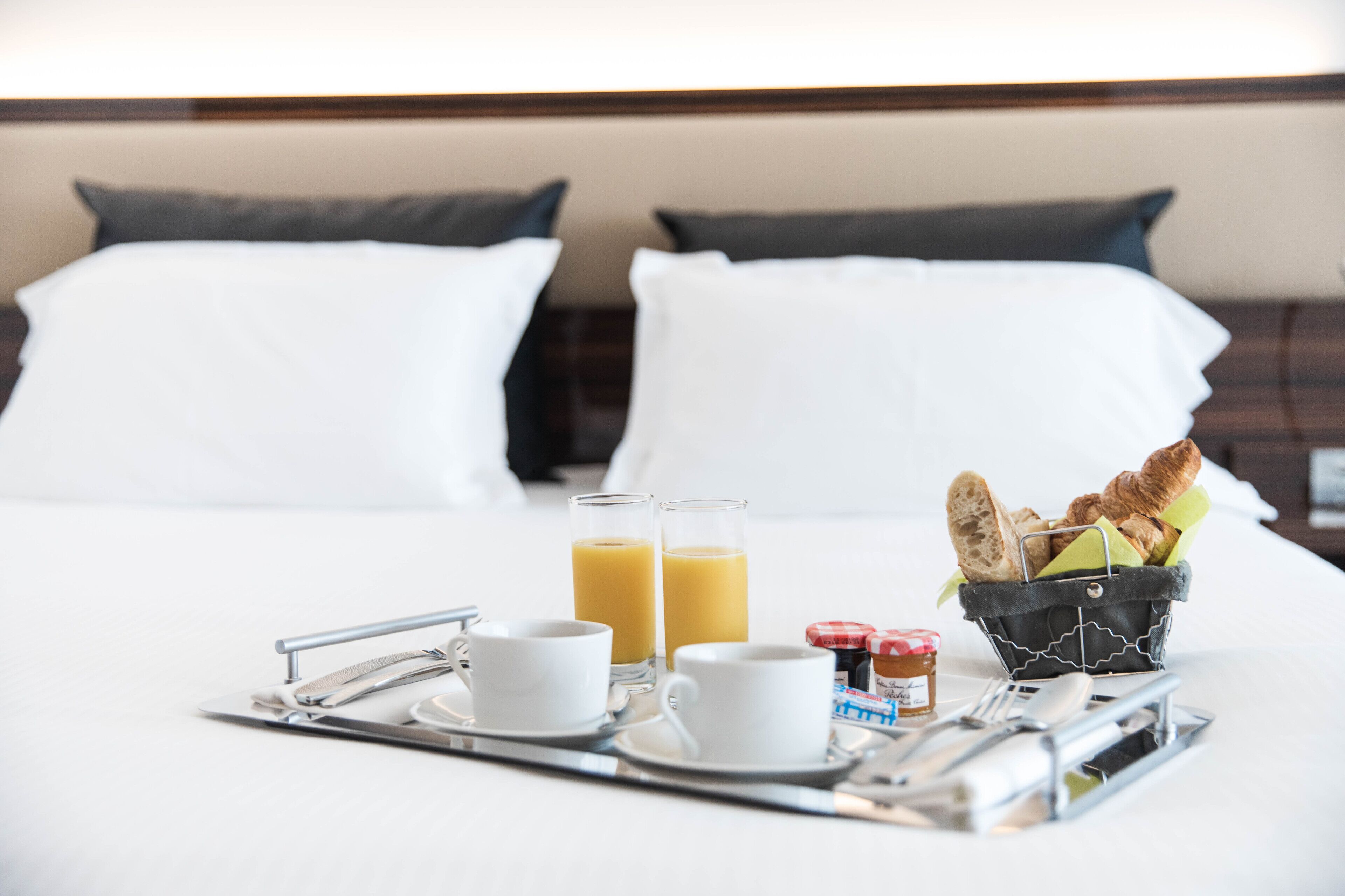 Ein schön gedecktes Frühstücksbrettchen mit zwei Orangensäften, zwei weißen Tassen, einem Marmeladenglas und einem Korb mit Croissants auf einem Hotelbett.