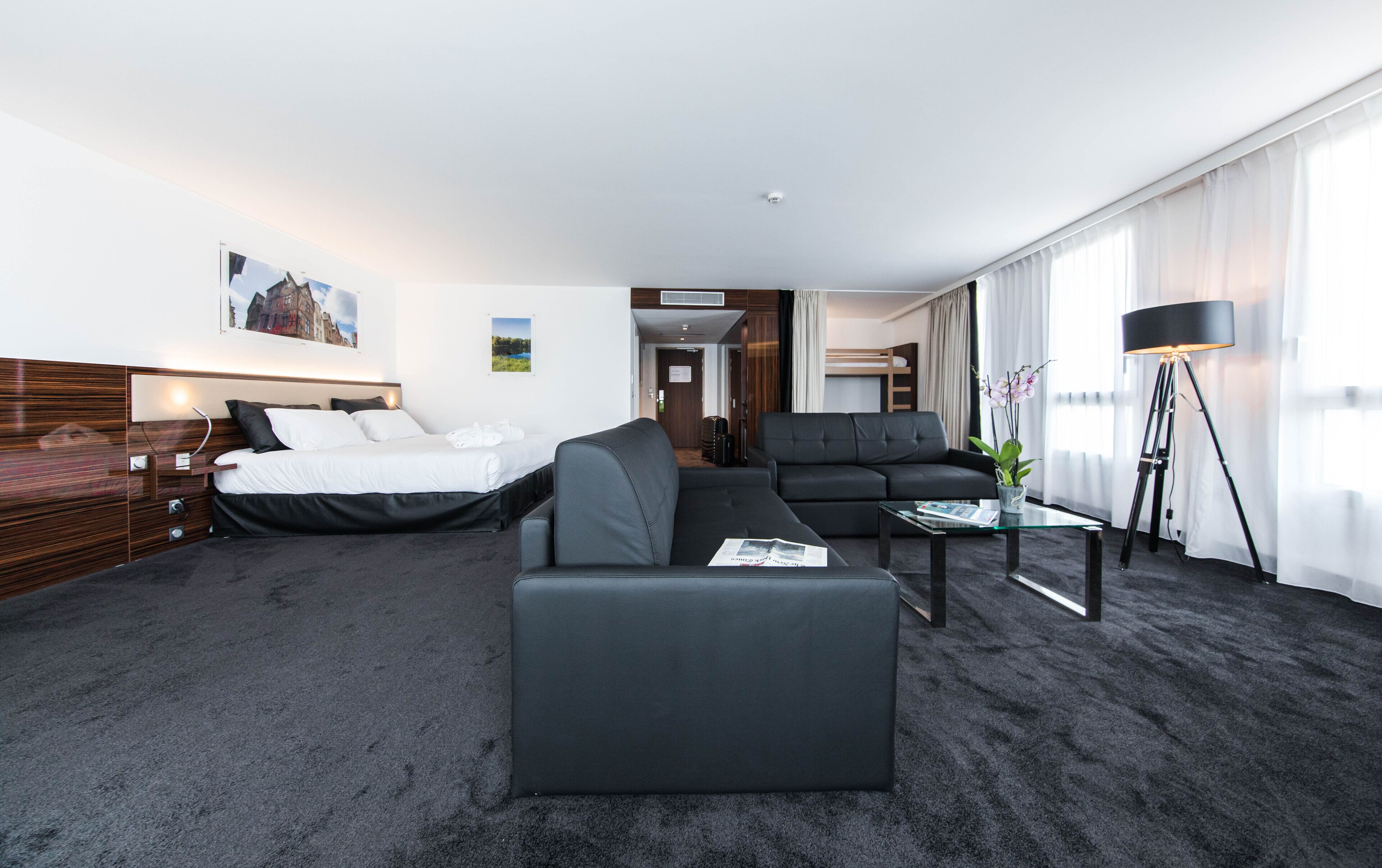 Habitació d'hotel moderna amb llit, sofà i butaca, una taula de cafè de vidre i una làmpada de peu.
