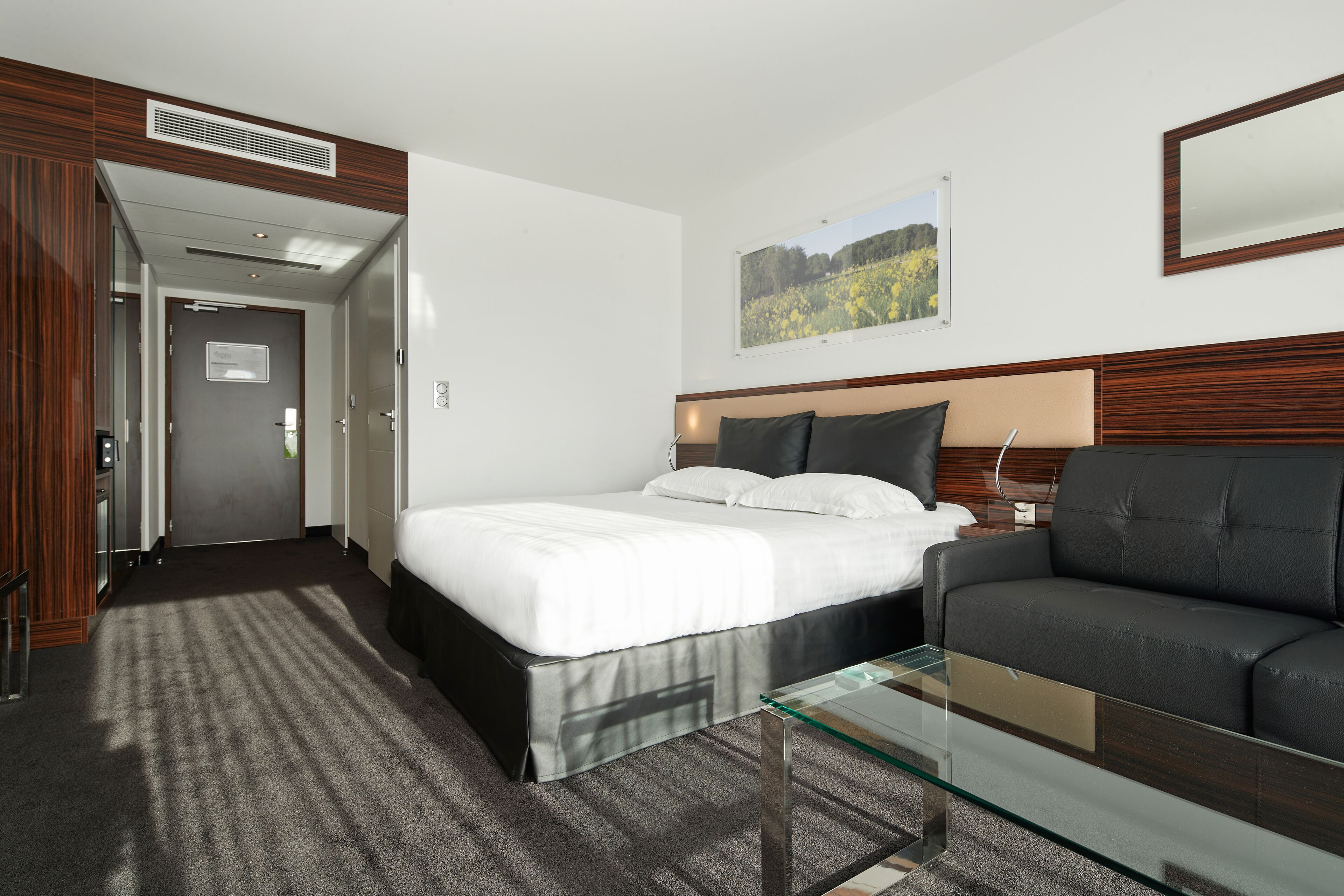 Une chambre d’hôtel comprenant un lit, une table basse en verre, un canapé, une tête de lit en bois, un tableau encadré au mur et une porte donnant sur une autre pièce.