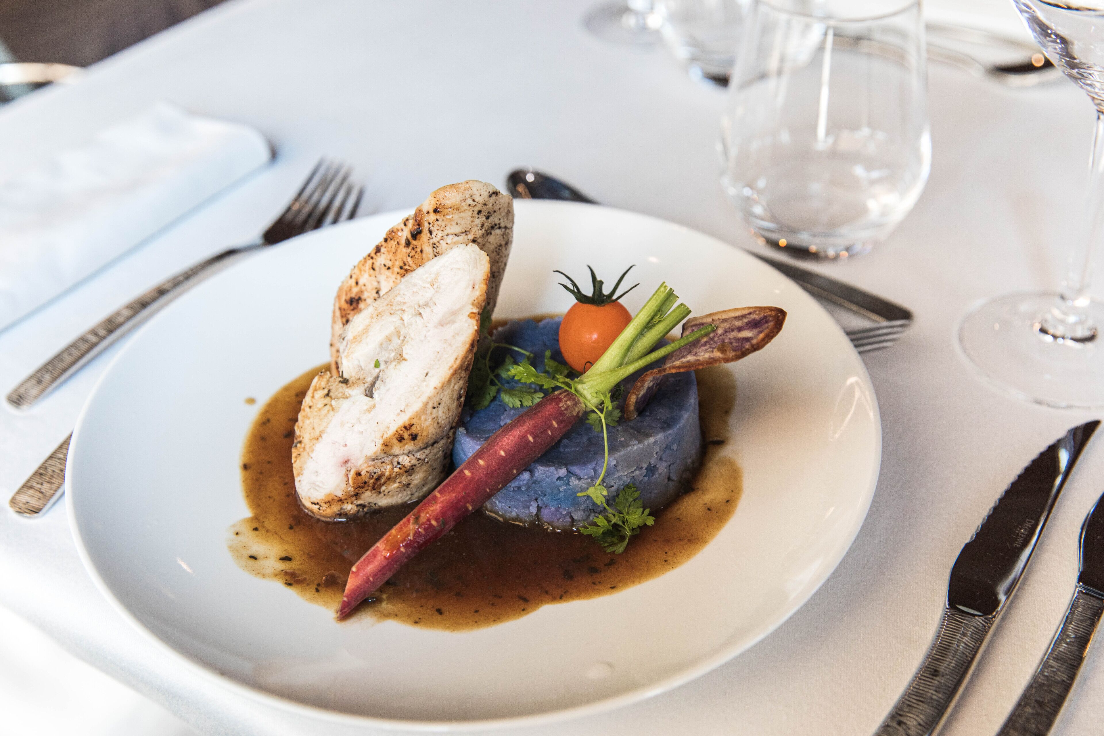 A white plate with a piece of grilled chicken, a carrot, and a small tomato on a blue puree, accompanied by a fork and knife on a white tablecloth.