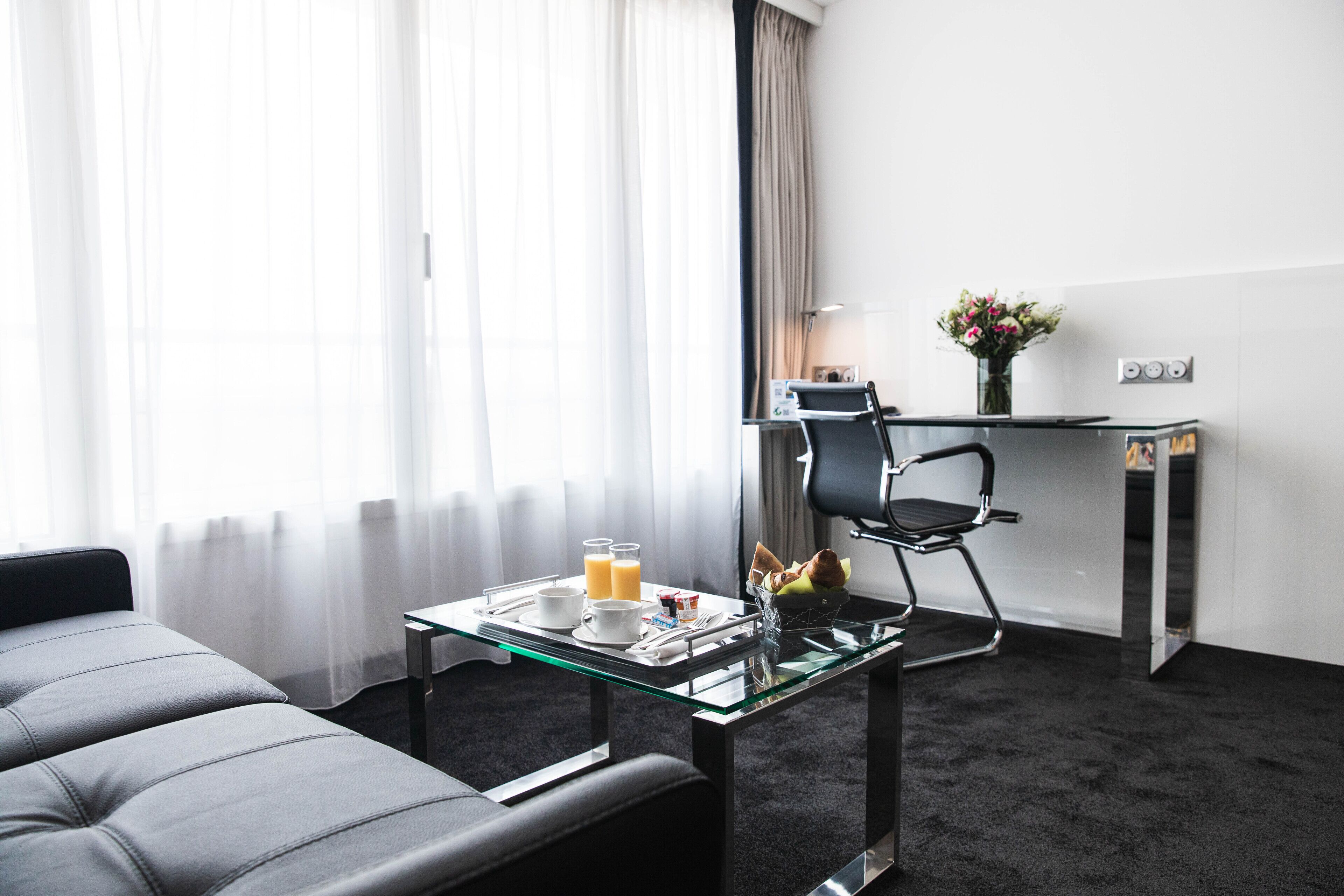 Una habitación de hotel moderna con una mesa de comedor de vidrio, una silla de oficina negra, un escritorio pequeño con una lámpara y una ventana con cortinas traslúcidas.