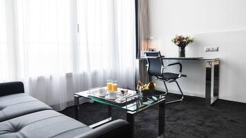 Una habitación de hotel moderna con una mesa de comedor de vidrio, una silla de oficina negra, un escritorio pequeño con una lámpara y una ventana con cortinas traslúcidas.