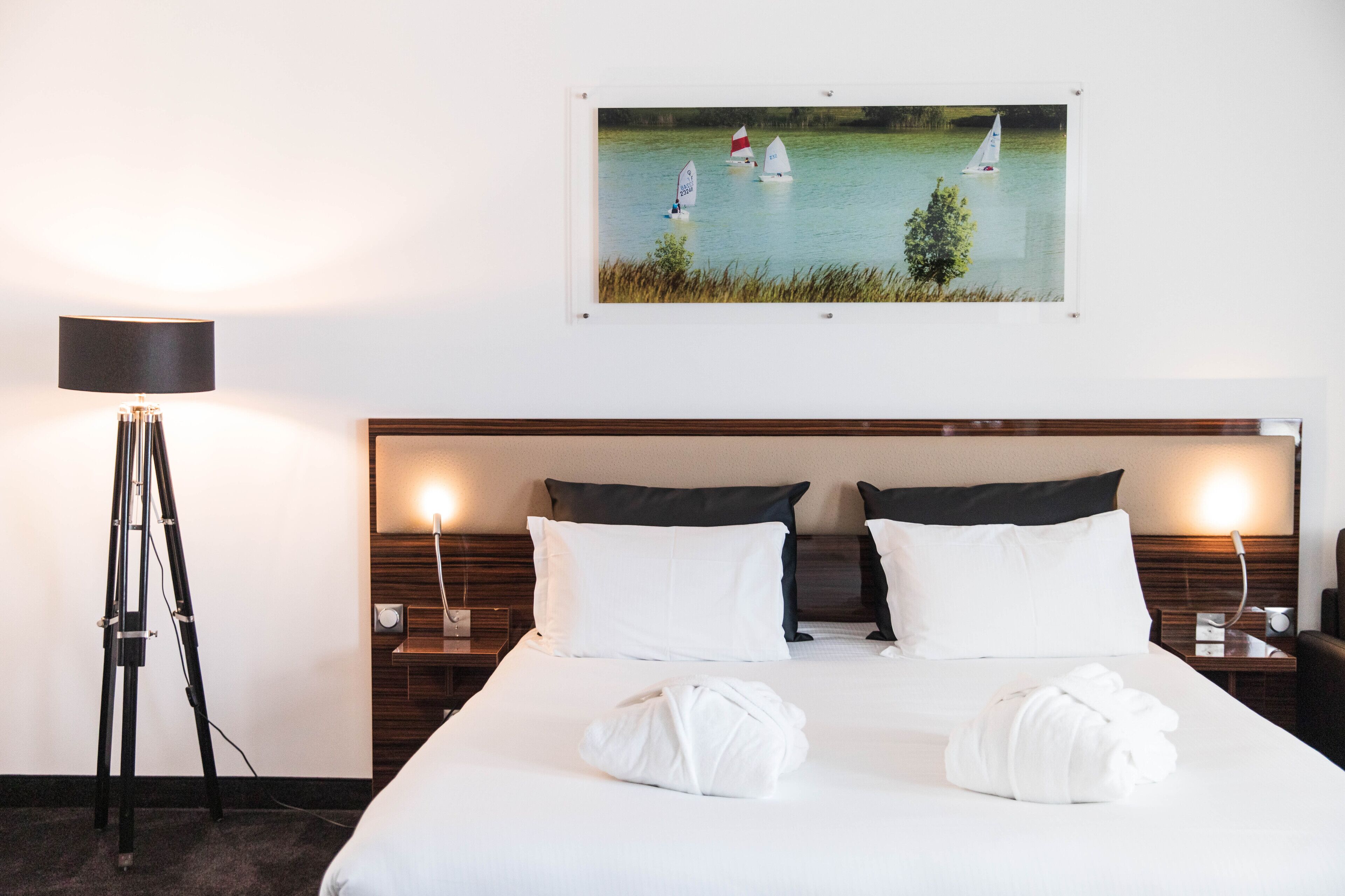 Habitación de hotel con una cama grande, dos almohadas, un cuadro enmarco con barcos de vela, una lámpara y un trípode.