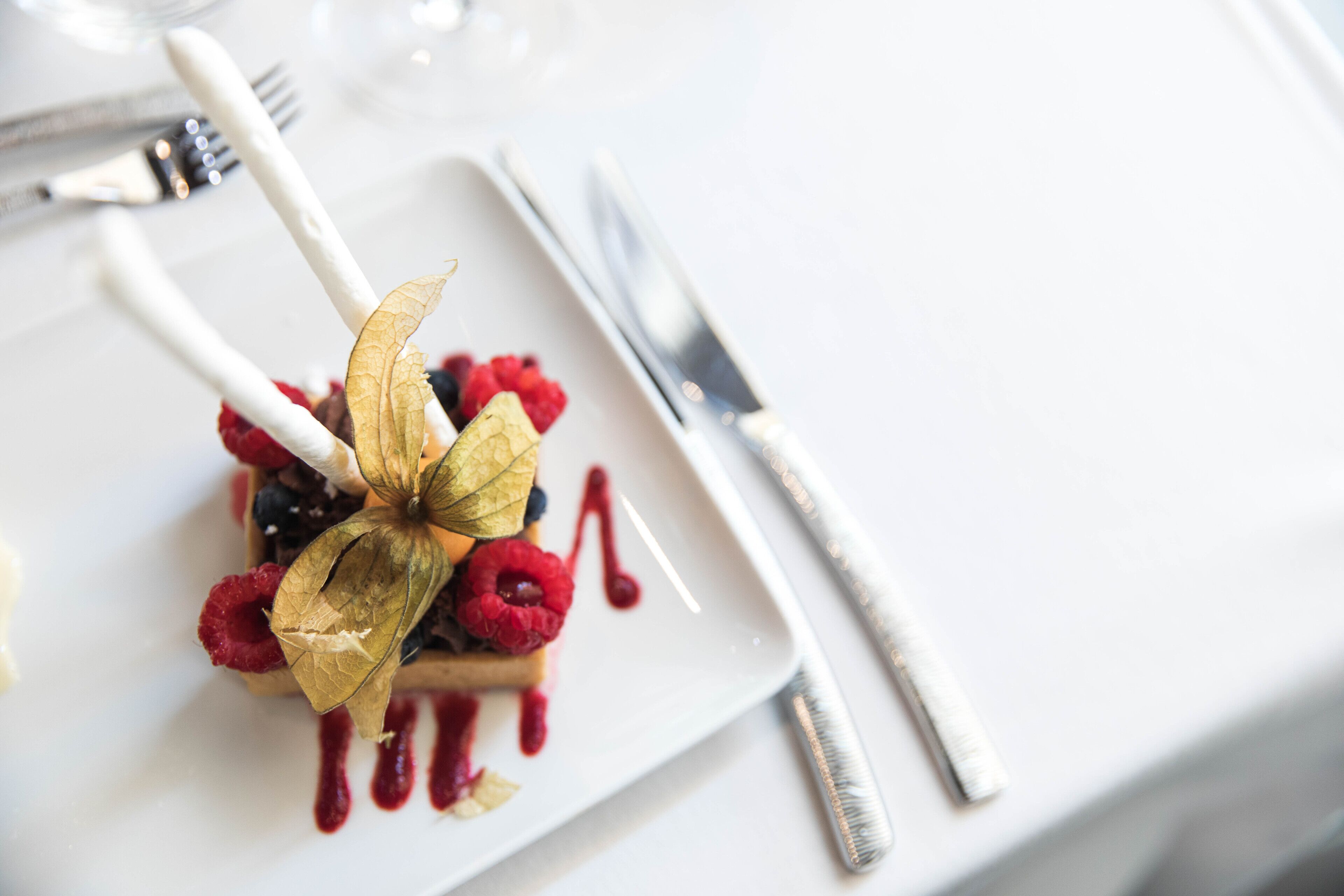 A dessert plate with berries and a decorative leaf, accompanied by a fork and knife on a white table.