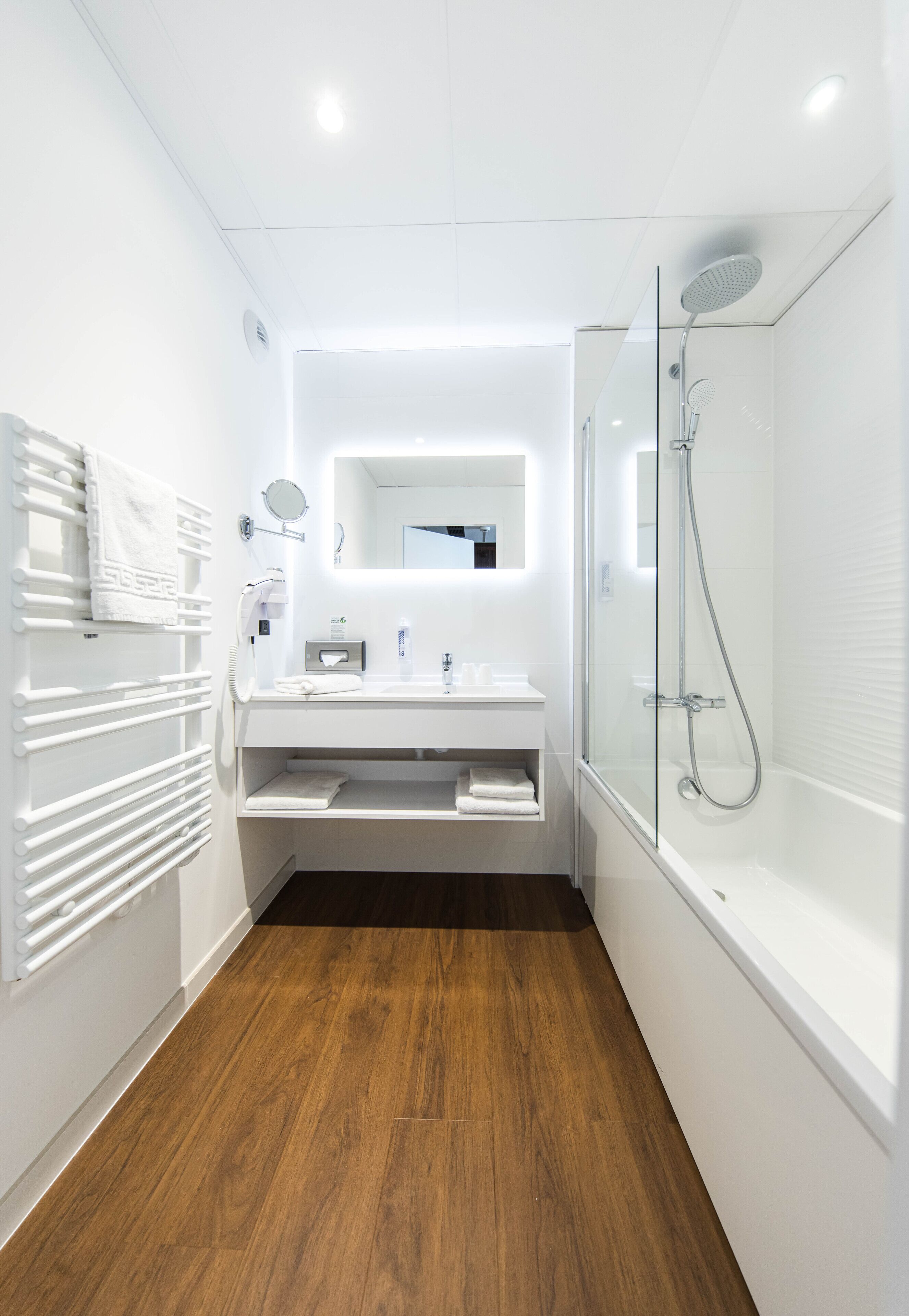 Une salle de bain moderne avec un plancher en bois, des accessoires blancs, une douche en verre et un meuble-lavabo intégré.
