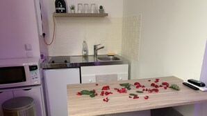 Uma cozinha compacta com micro-ondas, pia e pequena geladeira. Há pétalas de rosa sobre a mesa e um controle remoto ao lado.