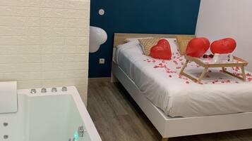 A modern bedroom with a large bathtub, a bed with heart-shaped pillows, and a small shelf with a plant.