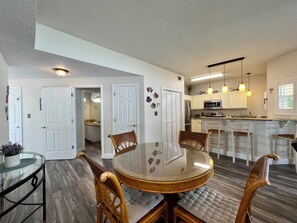 A modern kitchen with white cabinets, a stainless steel refrigerator, and a microwave. There is a dining area with a glass-top table and wicker chairs.