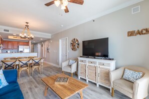 A spacious living area with a wooden coffee table, a TV on a cabinet, and a dining area with wooden chairs and a chandelier. - Seawind 1105 by Liquid Life (Gulf Shores)