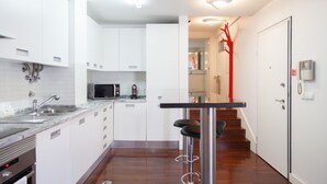 A modern kitchen with white cabinets, a stainless steel sink, and a microwave. There is a bar stool and a staircase leading to another room.