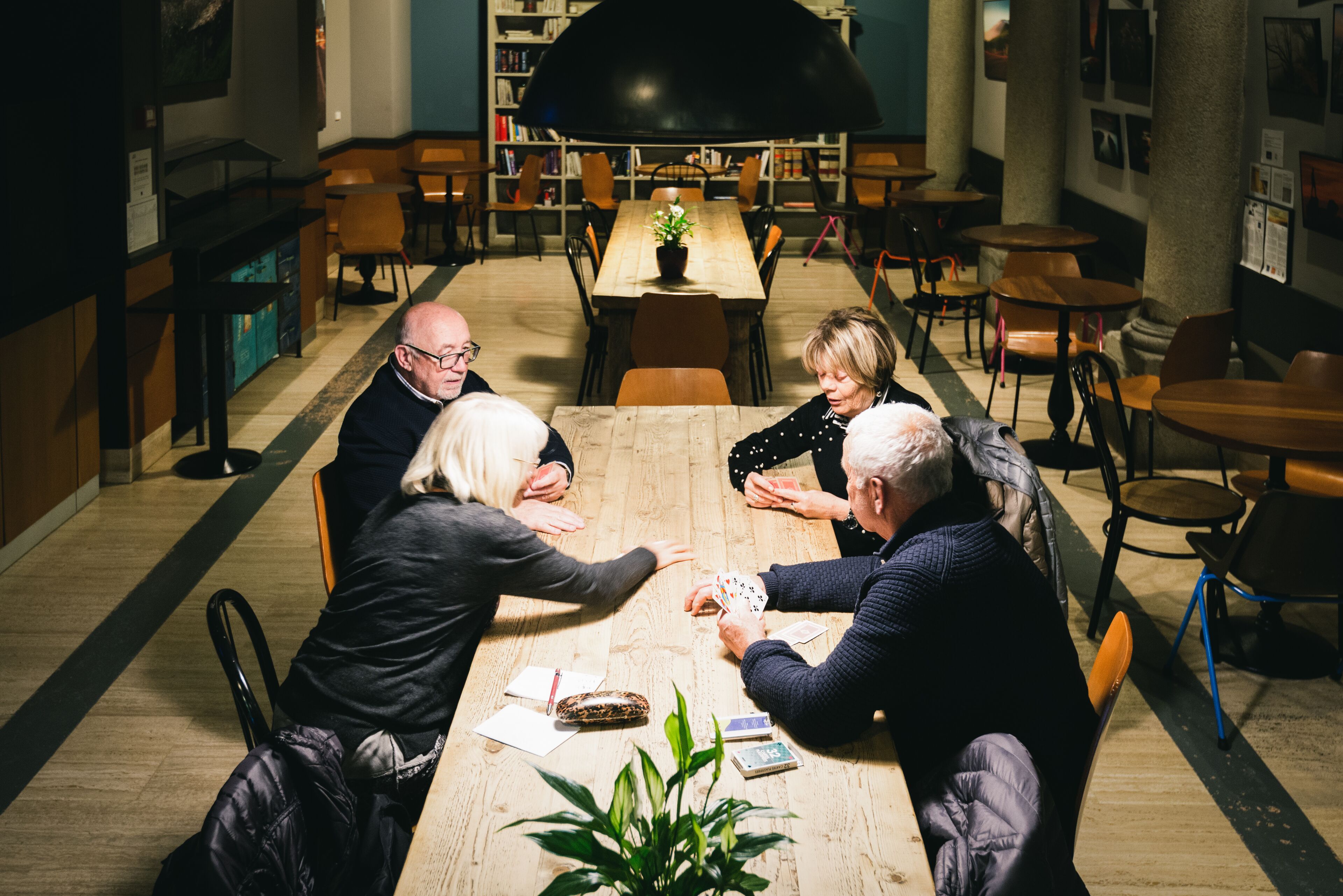 Un groupe de personnes joue aux cartes autour d’une table en bois, dans un espace intérieur bien éclairé, avec des étagères et des chaises en arrière-plan.