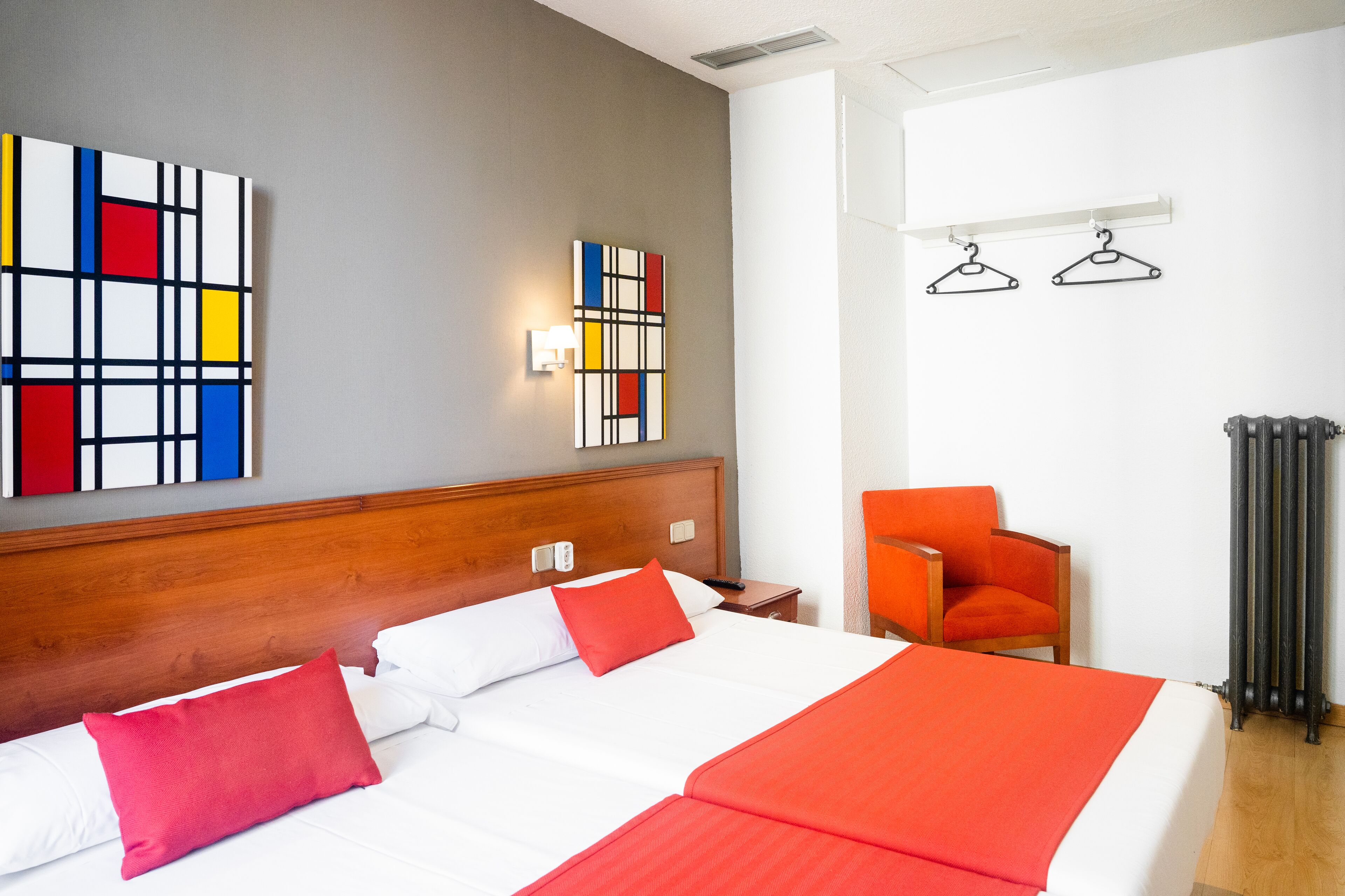 A hotel room with a wooden headboard, a bed with white and red bedding, a red chair, a radiator, and two abstract paintings on the wall.