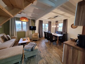 En rummelig opholdsstue med sofa, skammel, spisebord, stole og et fjernsyn.