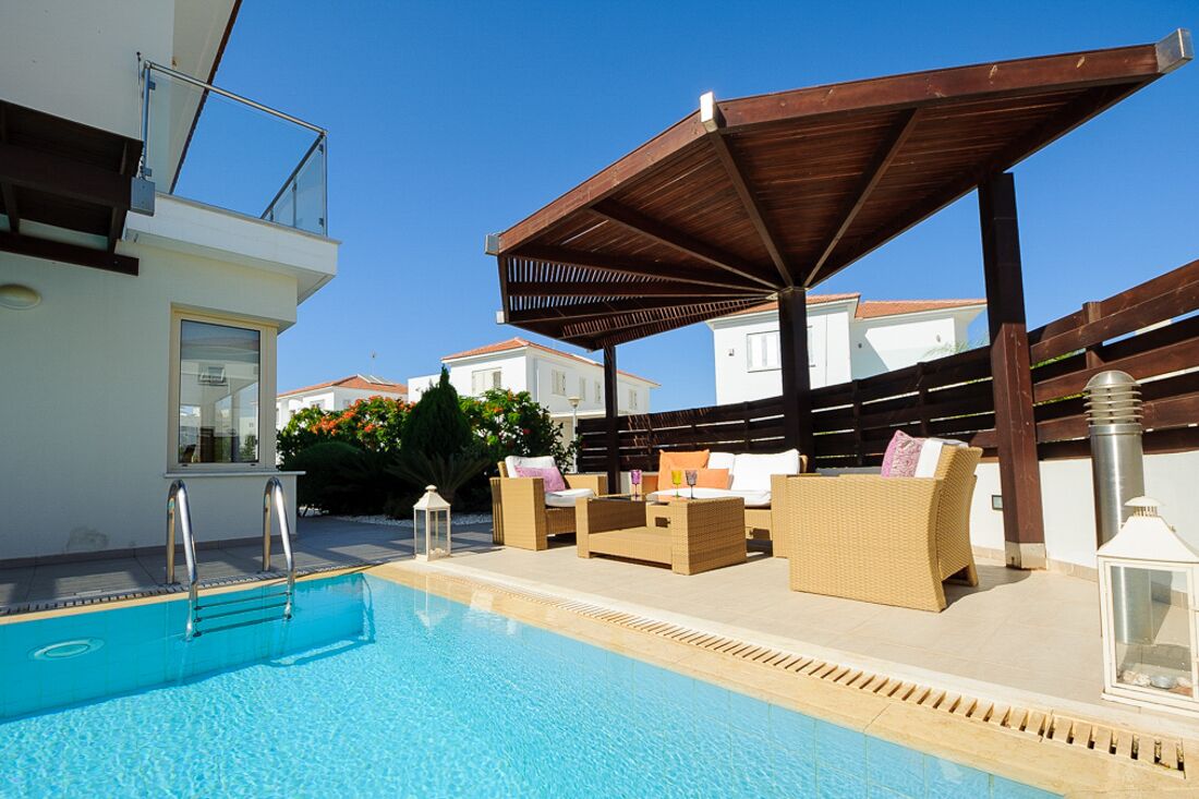 A pool area with a wooden pergola, lounge chairs, and a view of neighboring houses.