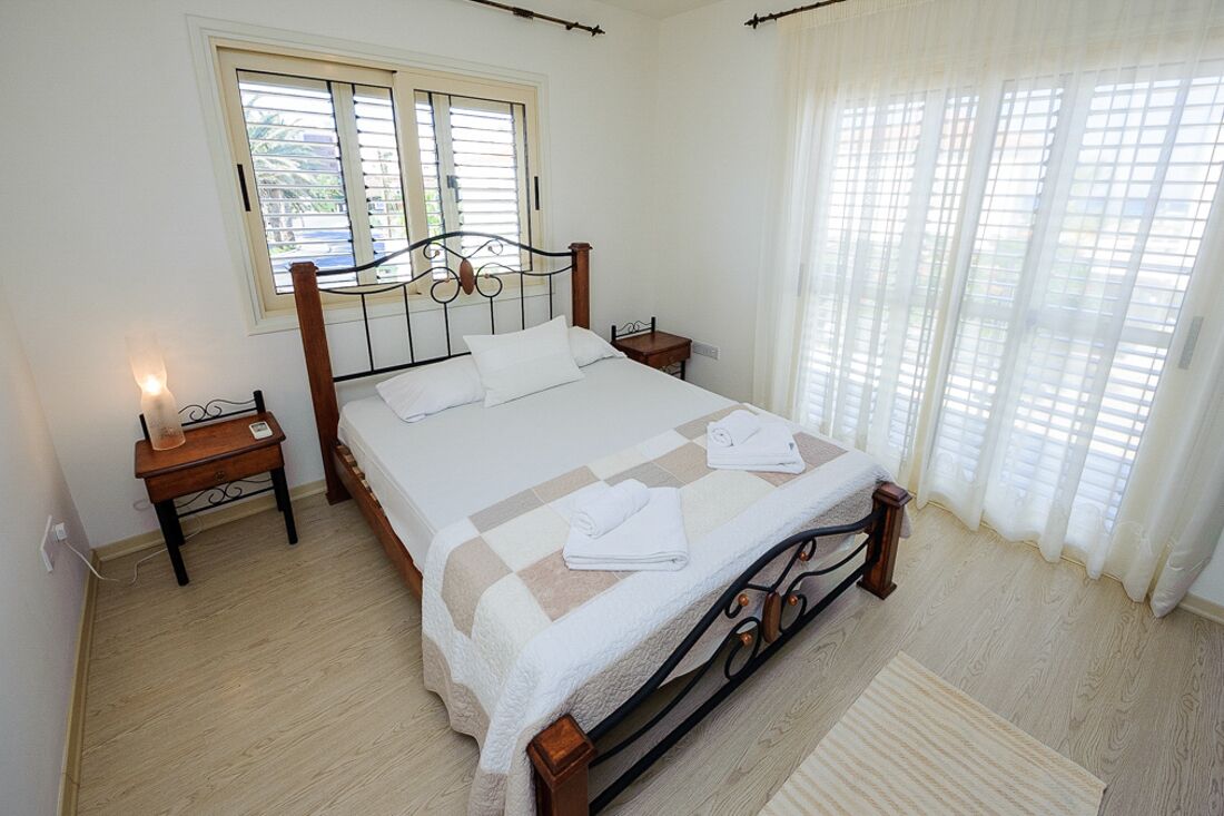 A neatly made bed with a checkered bedspread, a bedside table with a lamp, and a window with blinds.