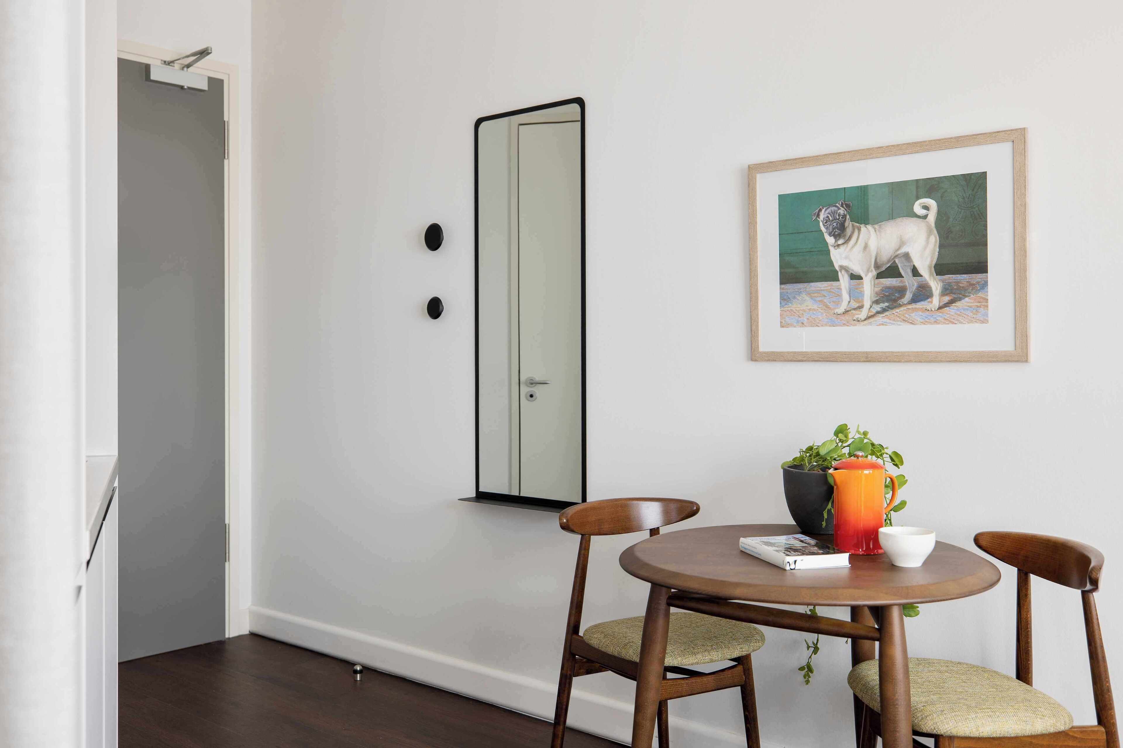 A dining area with a round table, two chairs, a framed dog painting, a potted plant, and a door with a mirror.