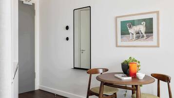 A dining area with a round table, two chairs, a framed dog painting, a potted plant, and a door with a mirror.