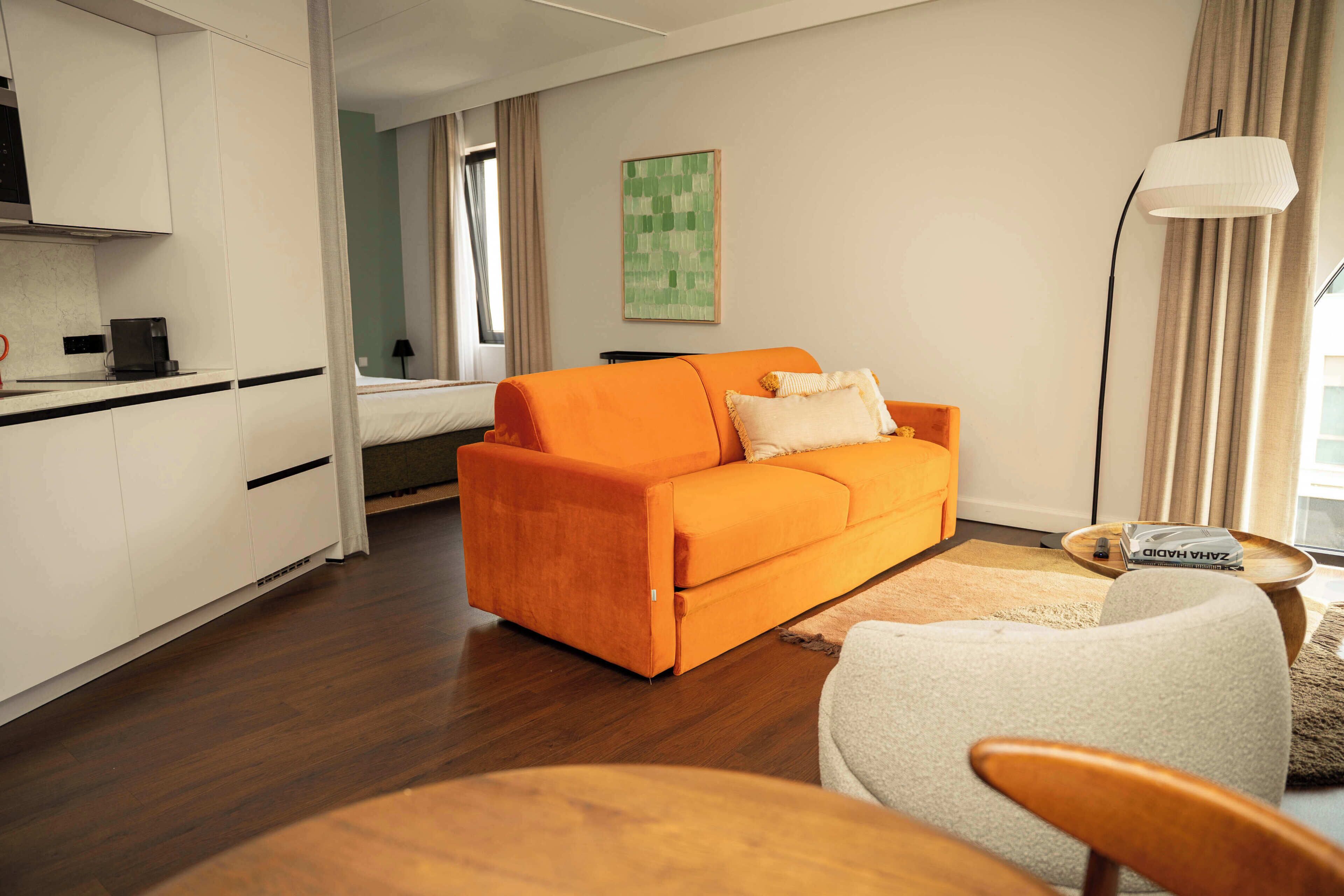 A modern hotel room with a bright orange sofa, a small round table, a white armchair, and a kitchen area with a coffee machine.