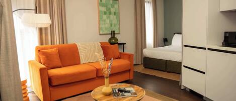 A modern hotel room with a sofa, a small wooden coffee table, a bed, and a kitchenette.