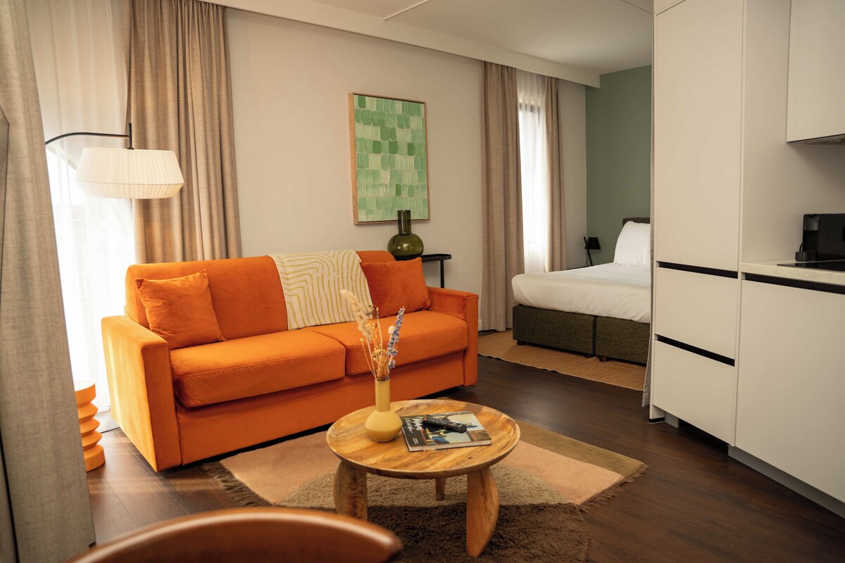 Ein modernes Hotelzimmer mit einer Couch, einem kleinen Holztisch, einem Bett und einer Küchenzeile.