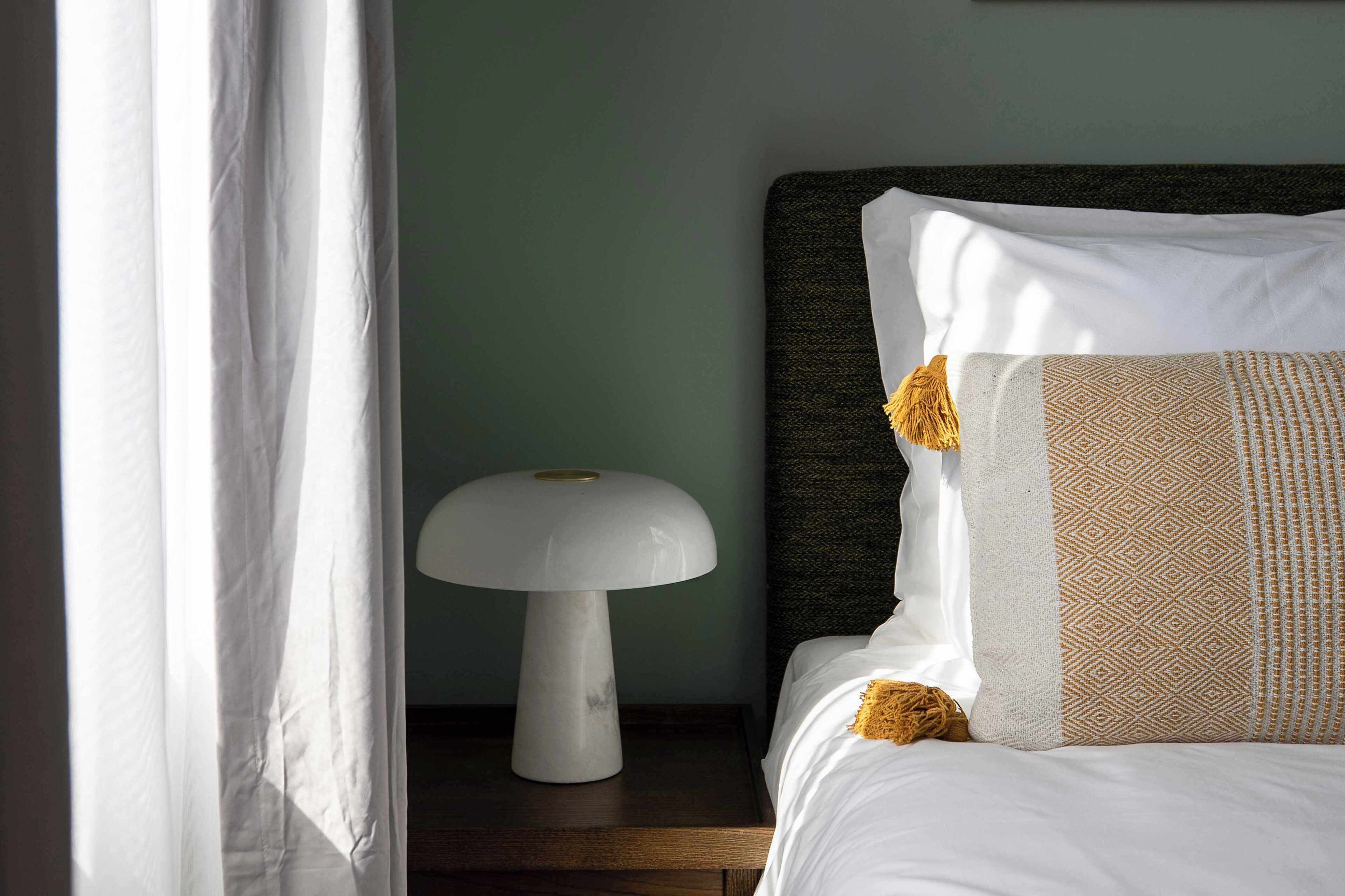 Un letto rifatto con cura, un cuscino decorato, una lampada bianca su un comodino in legno e una tenda leggera parzialmente scoperta.