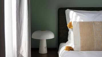 Un letto rifatto con cura, un cuscino decorato, una lampada bianca su un comodino in legno e una tenda leggera parzialmente scoperta.