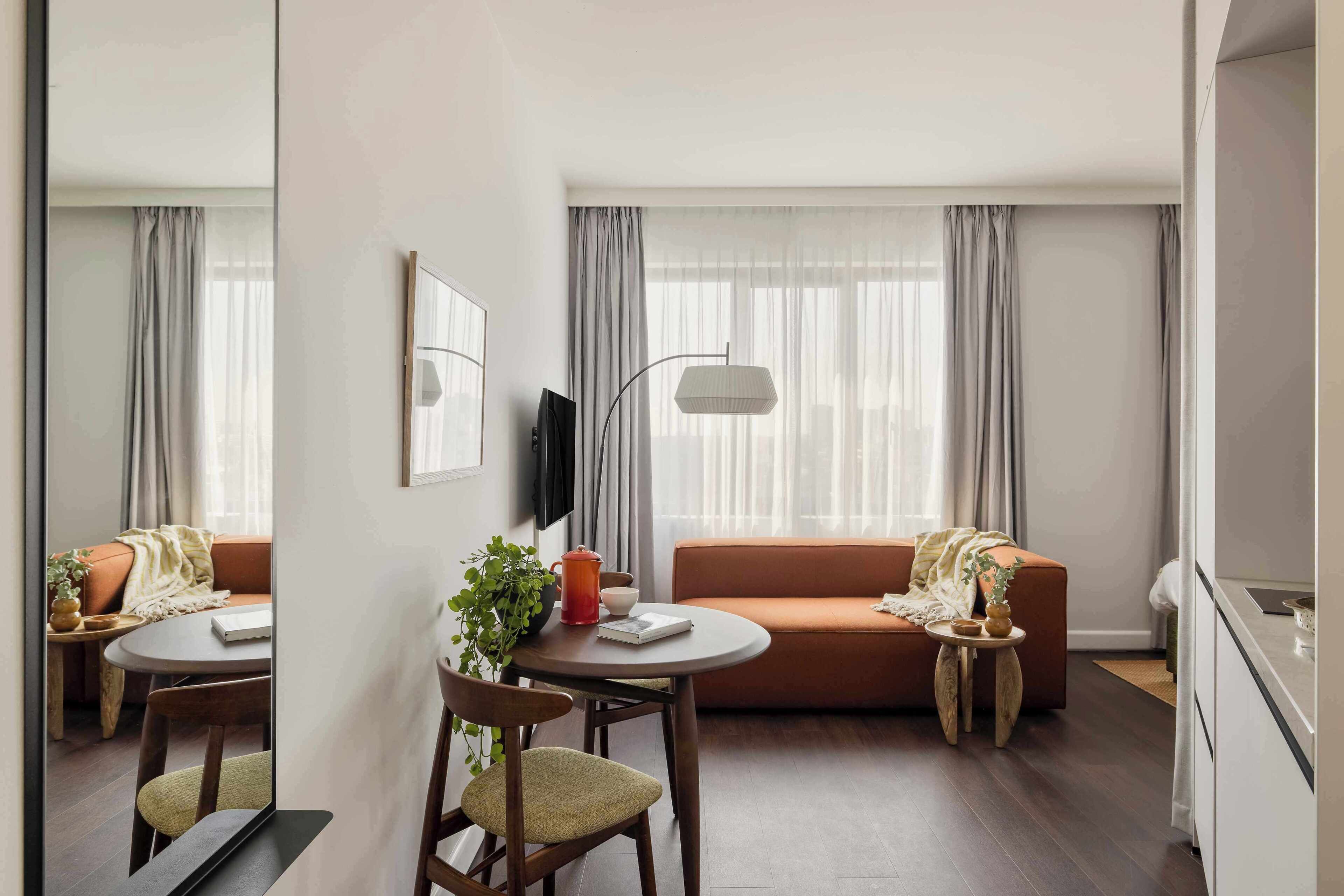 A modern living room with a sofa, a dining table with chairs, a TV mounted on the wall, and a large window with curtains.