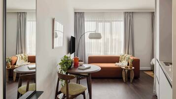 A modern living room with a sofa, a dining table with chairs, a TV mounted on the wall, and a large window with curtains.